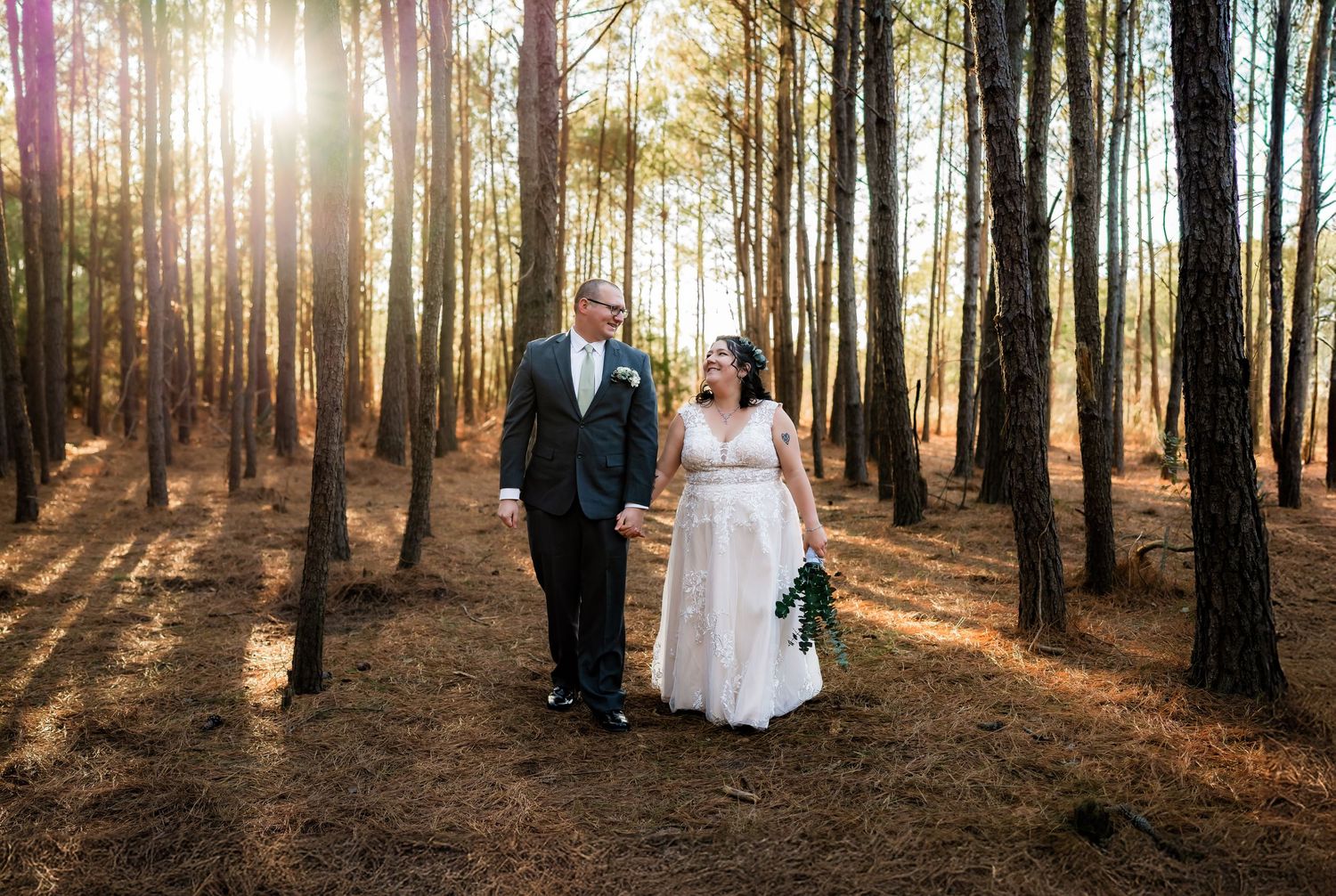 Couple walking hand in hand through sunlit forest in wedding attire.