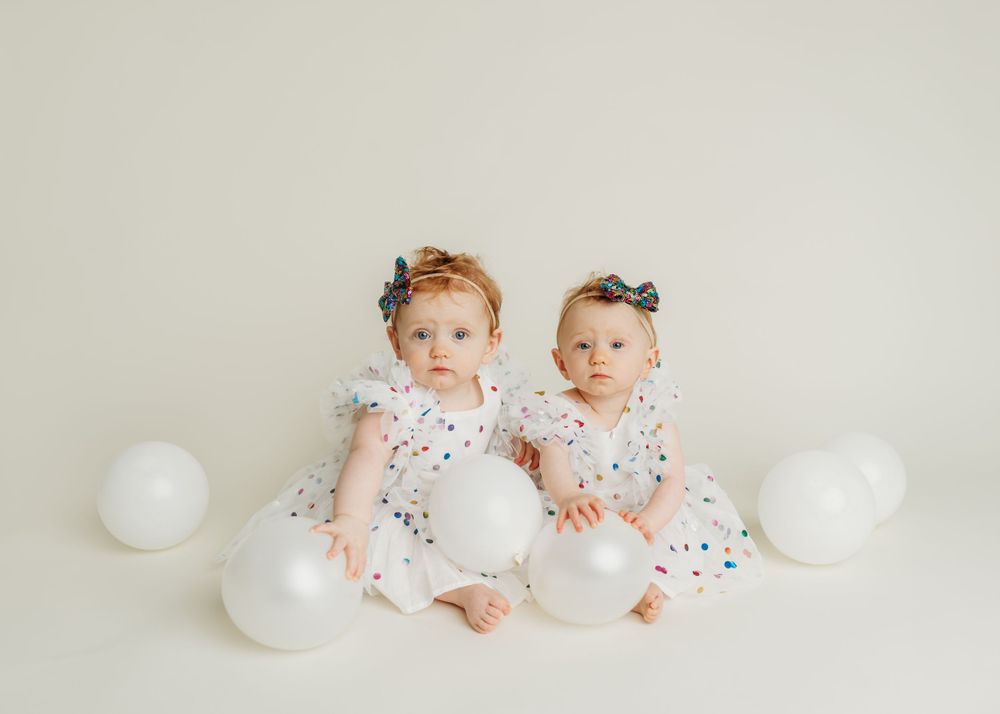 Two babies in polka dot outfits sitting together with white balloons.