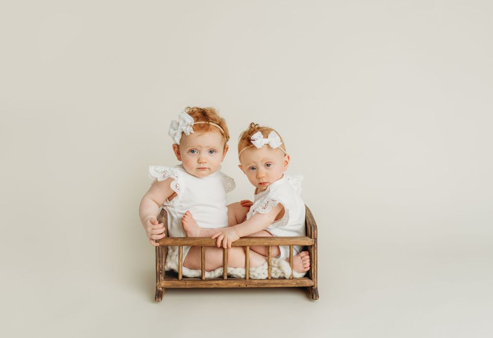 Two toddlers in white outfits sitting together in wooden bench.