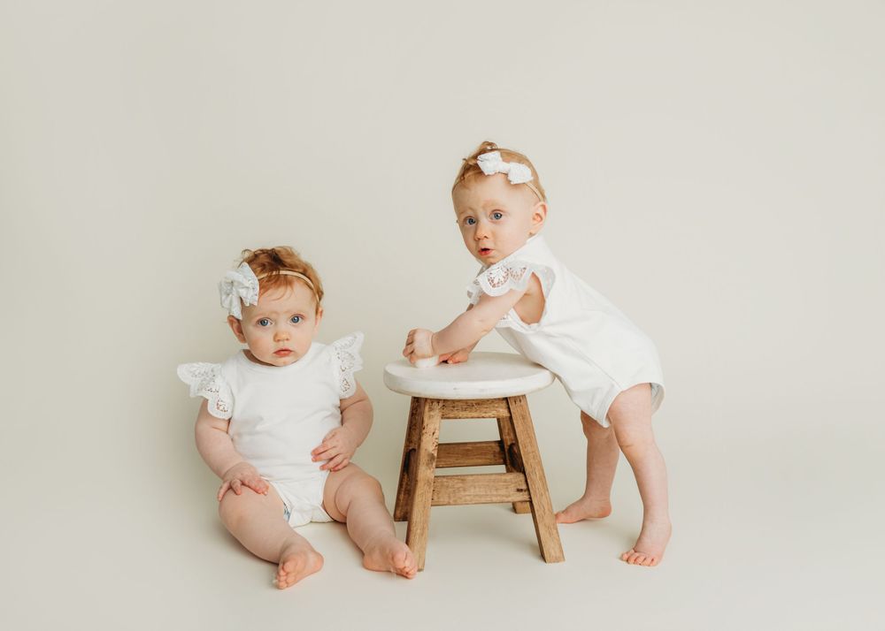 Two babies in white outfits pose with a wooden stool for a minimalist photo shoot.
