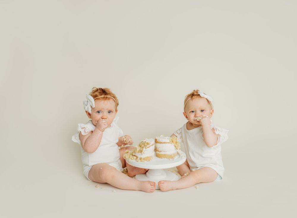 Two babies in white outfits sitting with a small cake between them.