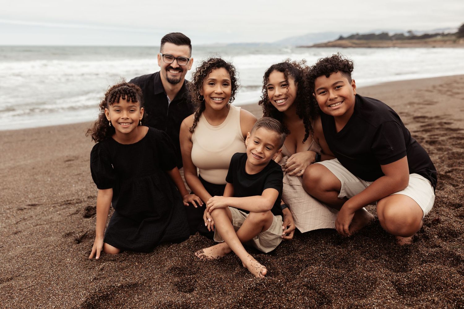 family of 6 sitting on a golden beach on a cloudy but bright day, all smiling