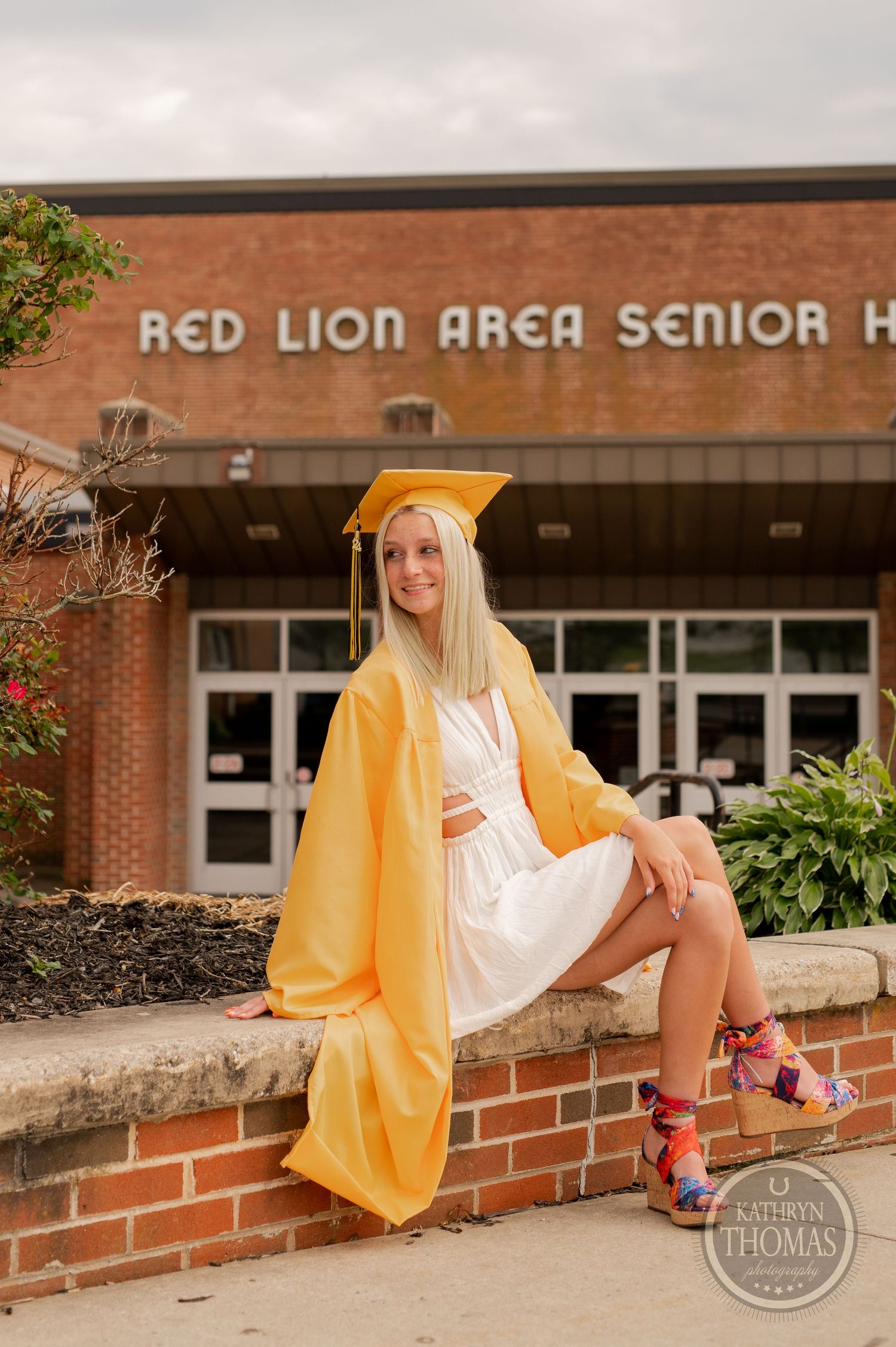 Graduate in white dress and yellow gown sits on brick wall outside high school entrance.
