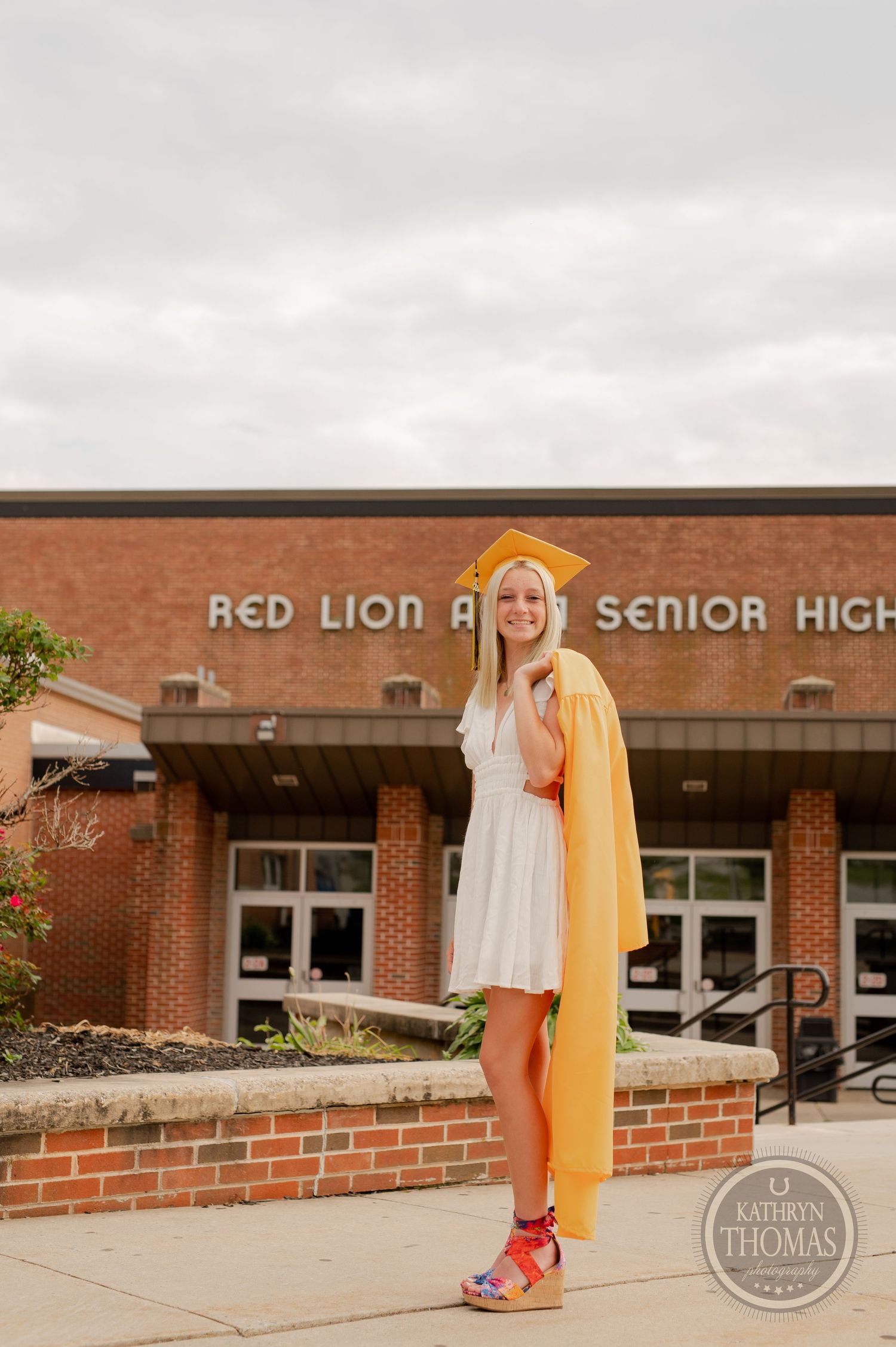 Student wearing a white dress and yellow cape stands on brick ledge at Red Lion High School.