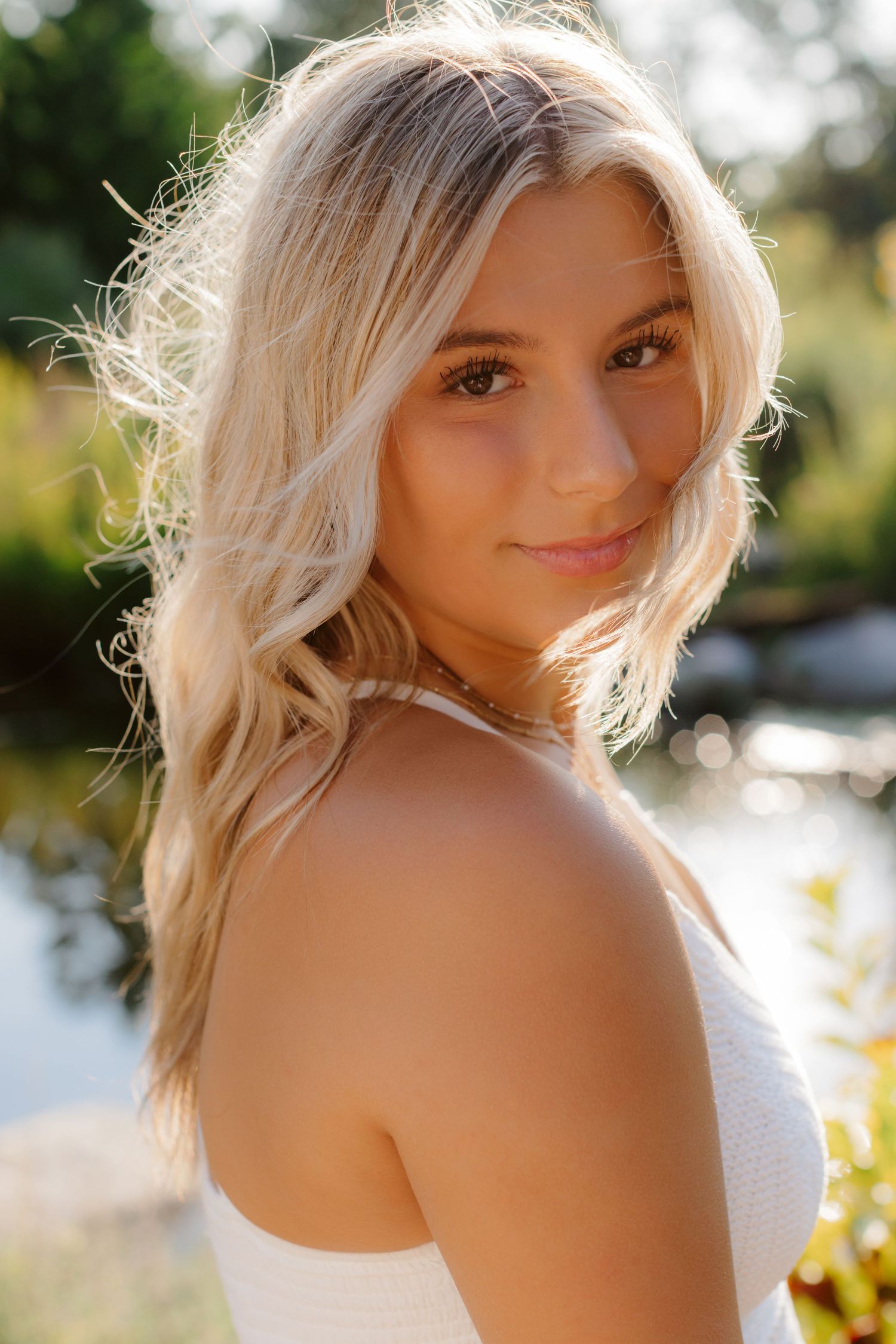 Blonde woman in white top poses outdoors with natural backlit glow in garden setting.