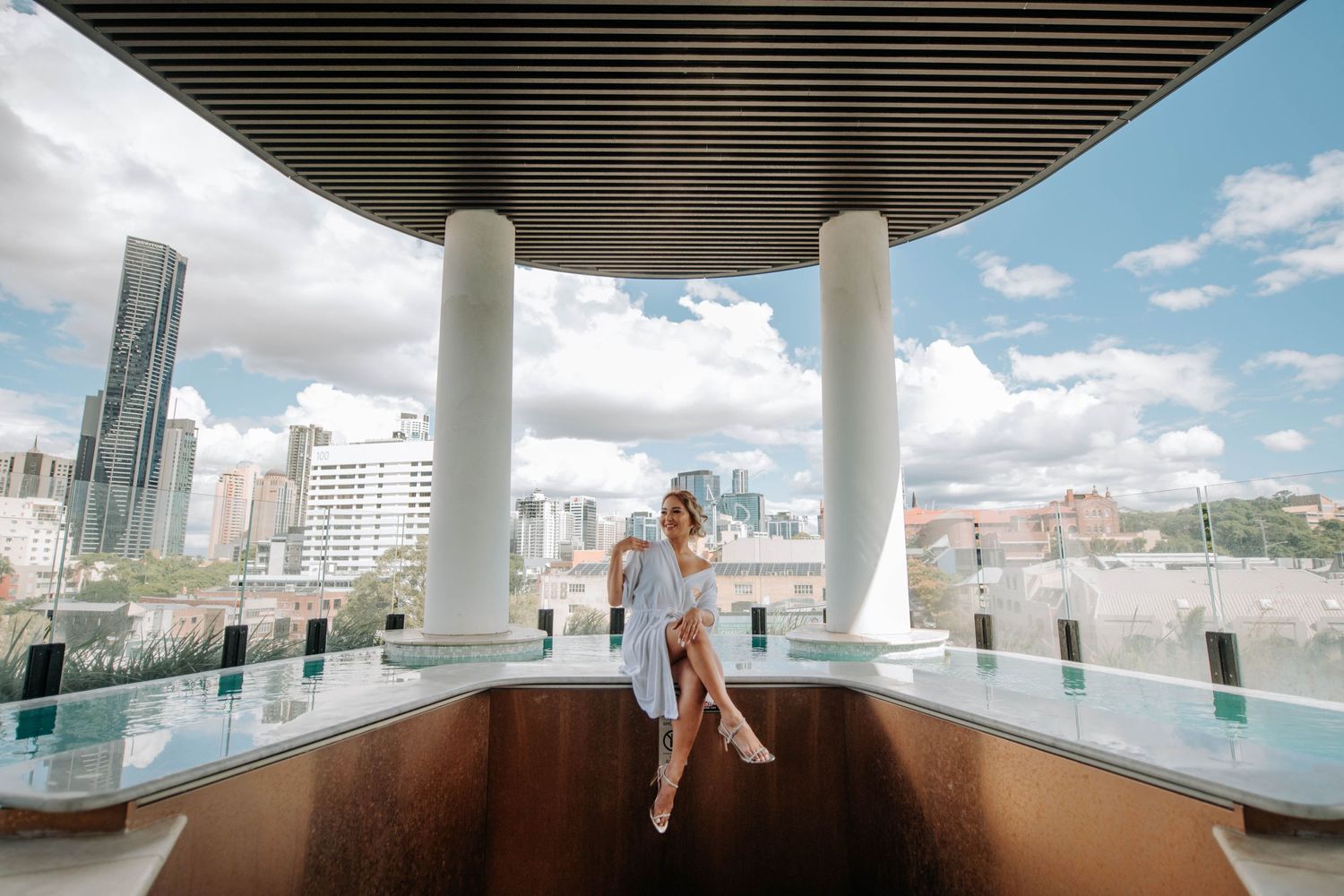Elegant figure sits on a balcony ledge with white columns and city skyline views on a sunny day.