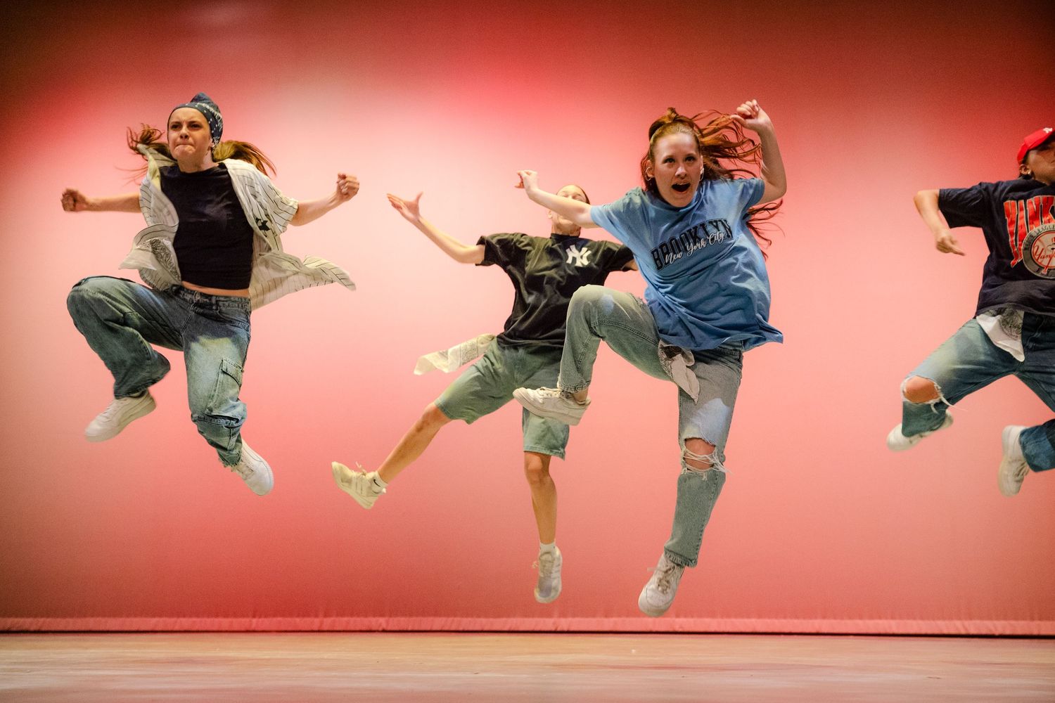 Group of dancers leaping simultaneously against coral colored wall in casual attire.
