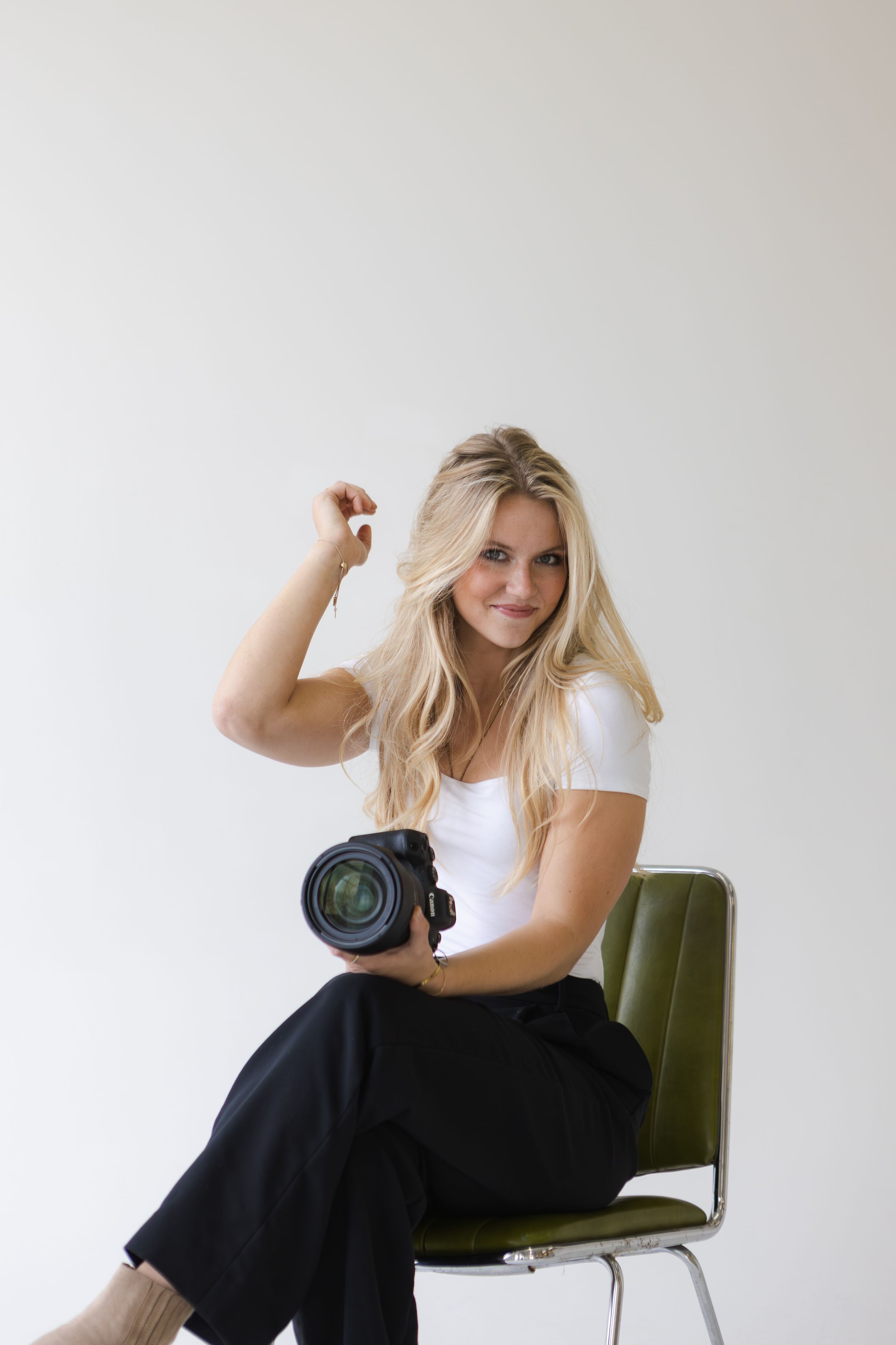 A photographer in white shirt sits on green chair holding professional camera in bright studio setting.