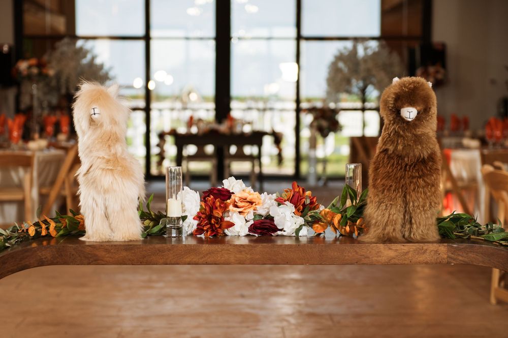 Two decorative plush alpacas flank a rustic wooden table adorned with floral arrangements and event decor.
