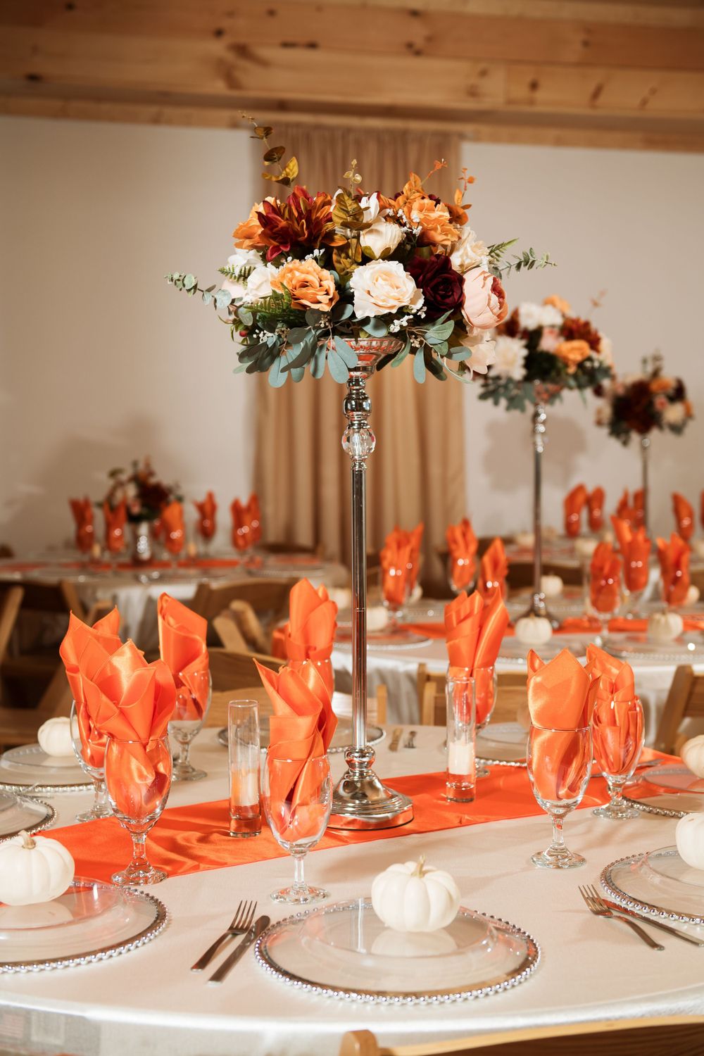 Elegant wedding table setup with tall floral centerpieces, orange napkins, and white place settings at a rustic venue.