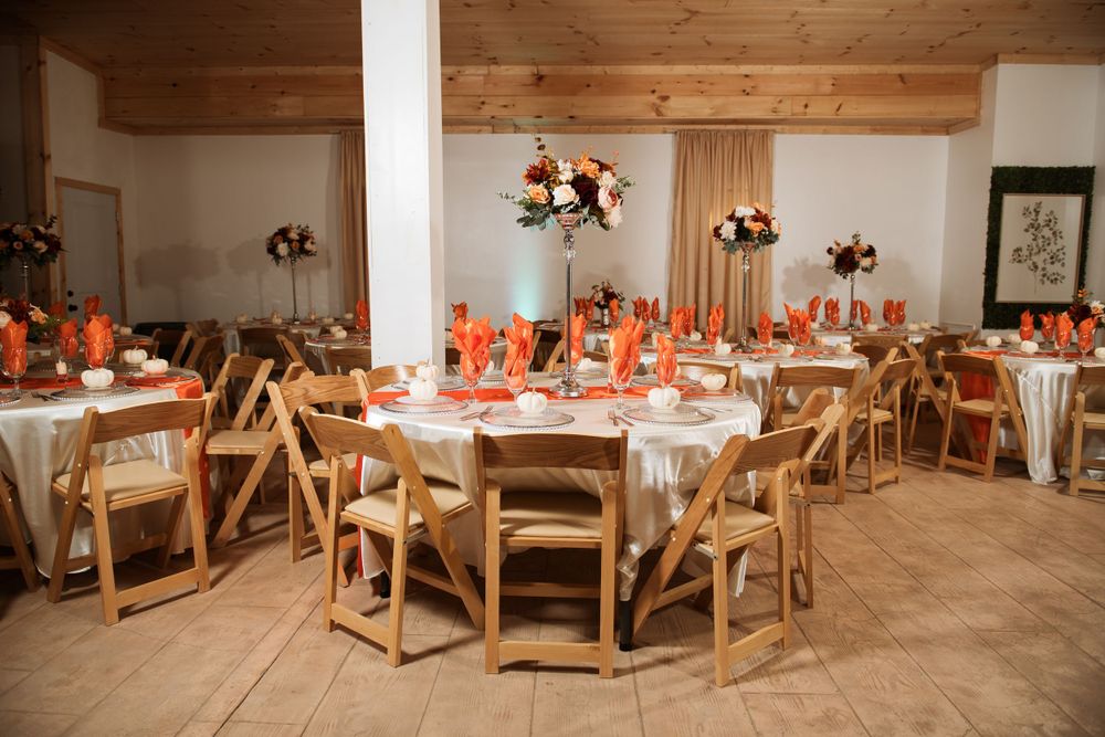 Wedding reception hall with round tables, wooden chairs, and orange accents under exposed wood beams.