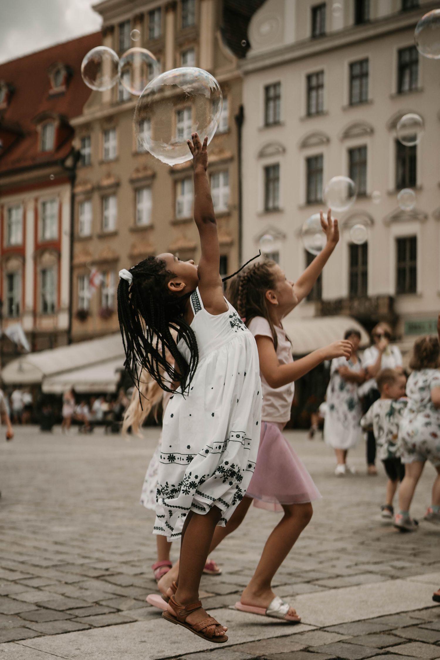 Two children in summer dresses jumping and reaching for bubbles in front of historic European buildings.