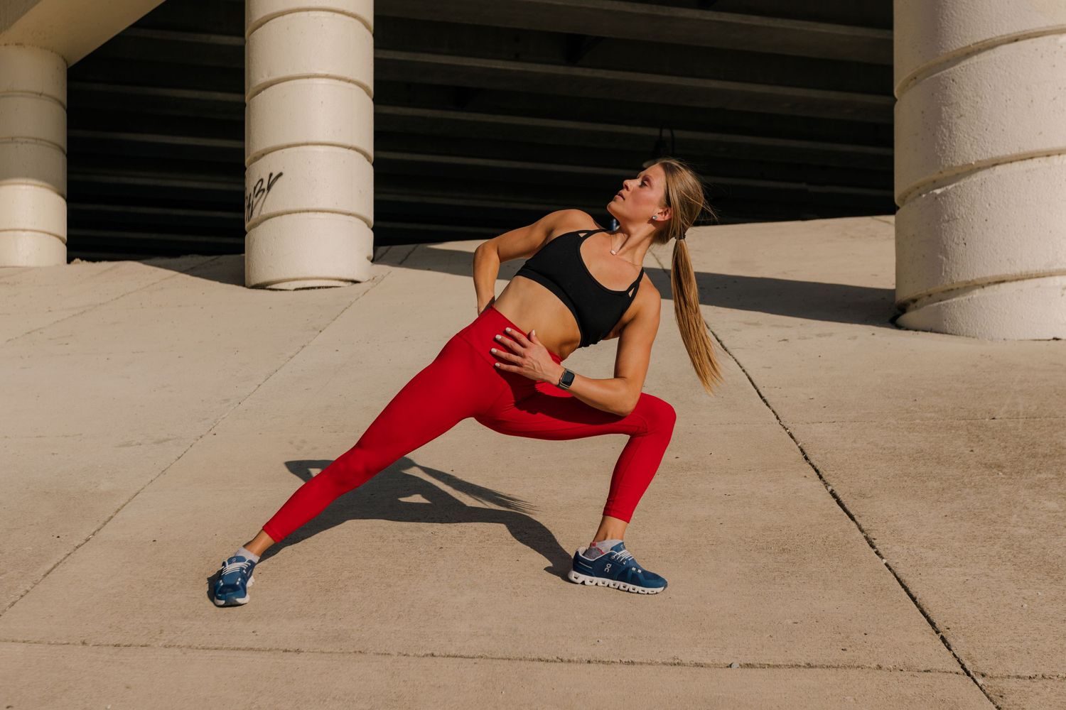 Athletic figure in red leggings performs a deep stretching lunge pose against an architectural backdrop with columns.