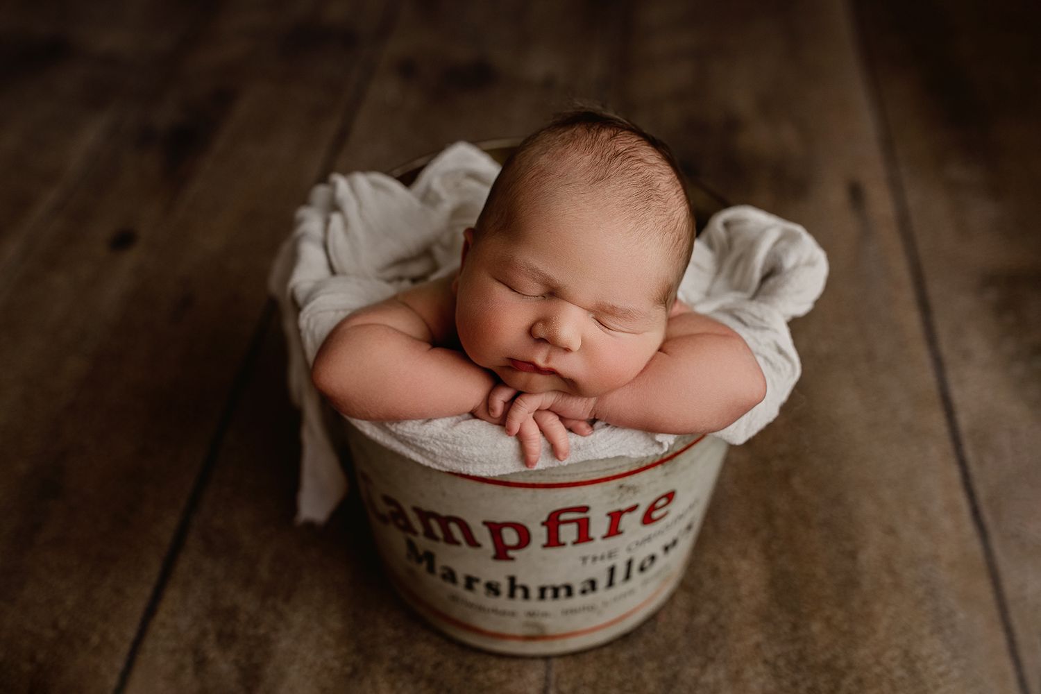 Sleeping newborn nestled in cozy white fabric inside a rustic wooden bowl on dark wooden surface.
