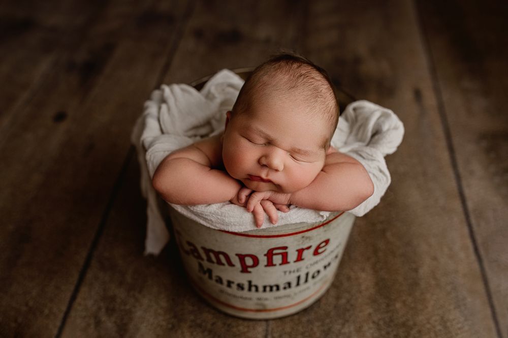 Sleeping newborn nestled in cozy white fabric inside a rustic wooden bowl on dark wooden surface.