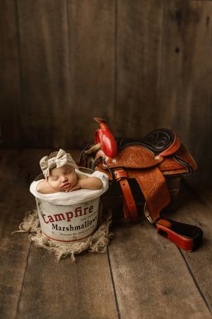 Artistic newborn photography series with western saddle and vintage marshmallow tin on rustic wooden backdrop.