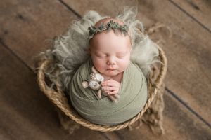 Sleeping newborn wrapped in sage green swaddle rests in rustic basket with floral headpiece on wooden floor.