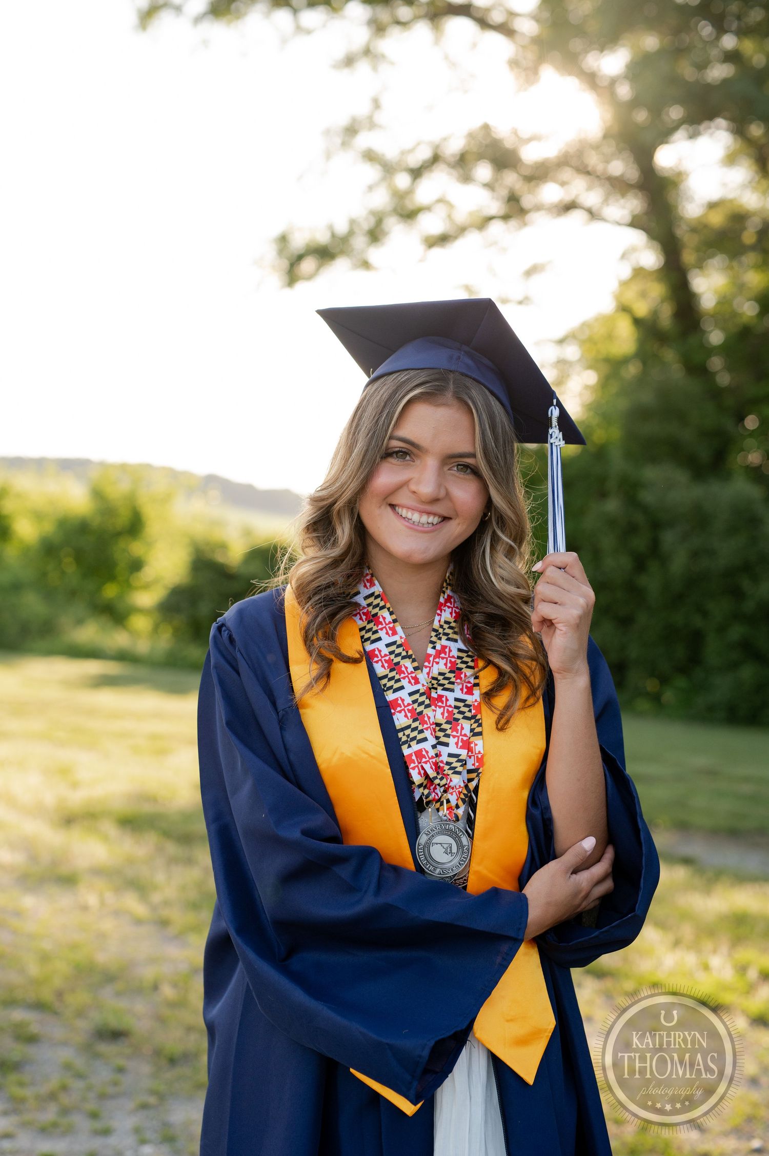 Graduate in academic regalia holding tassel while posing in natural outdoor setting.