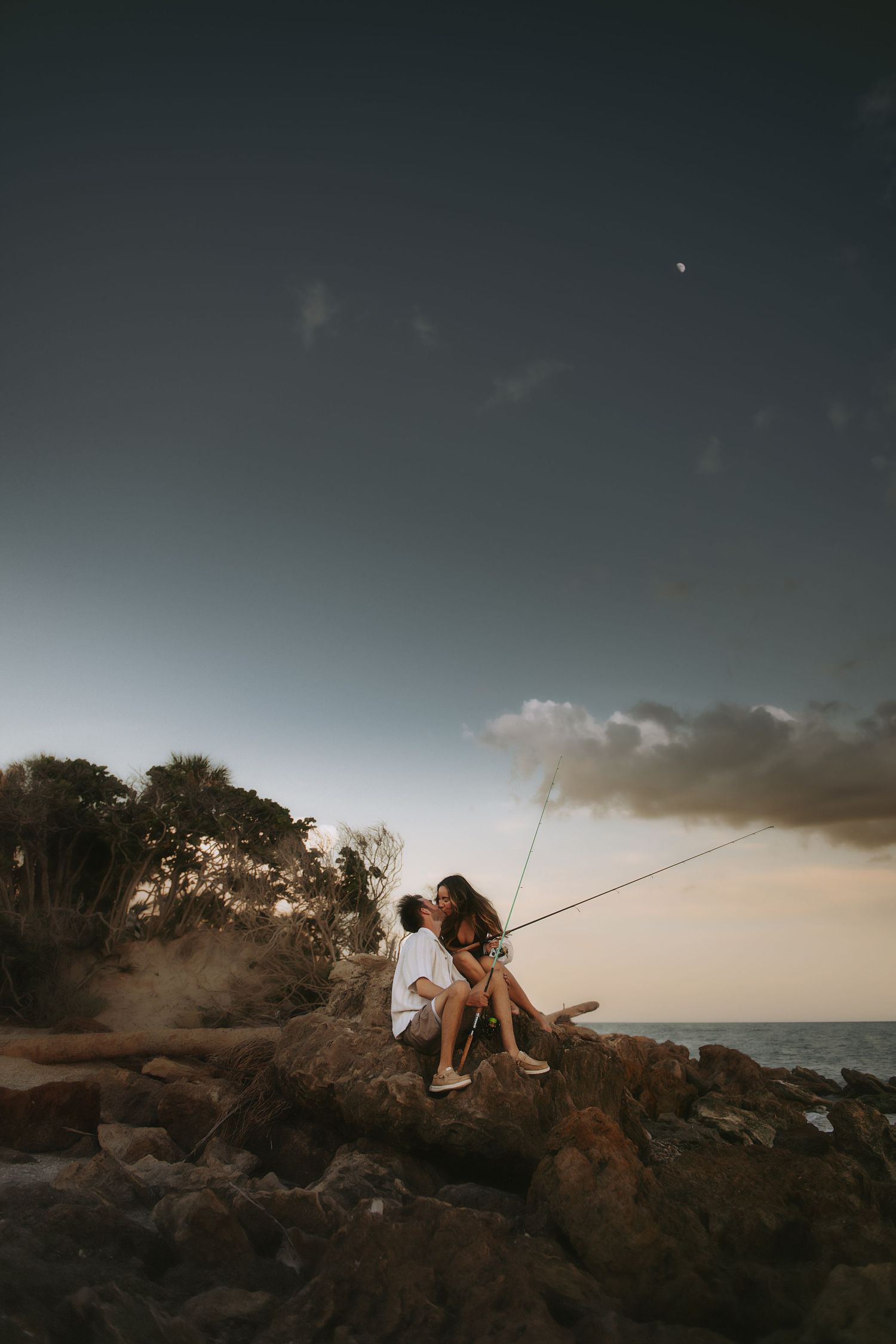 Couple fishing together on rocky coastal outcrop at dusk with moon in sky.