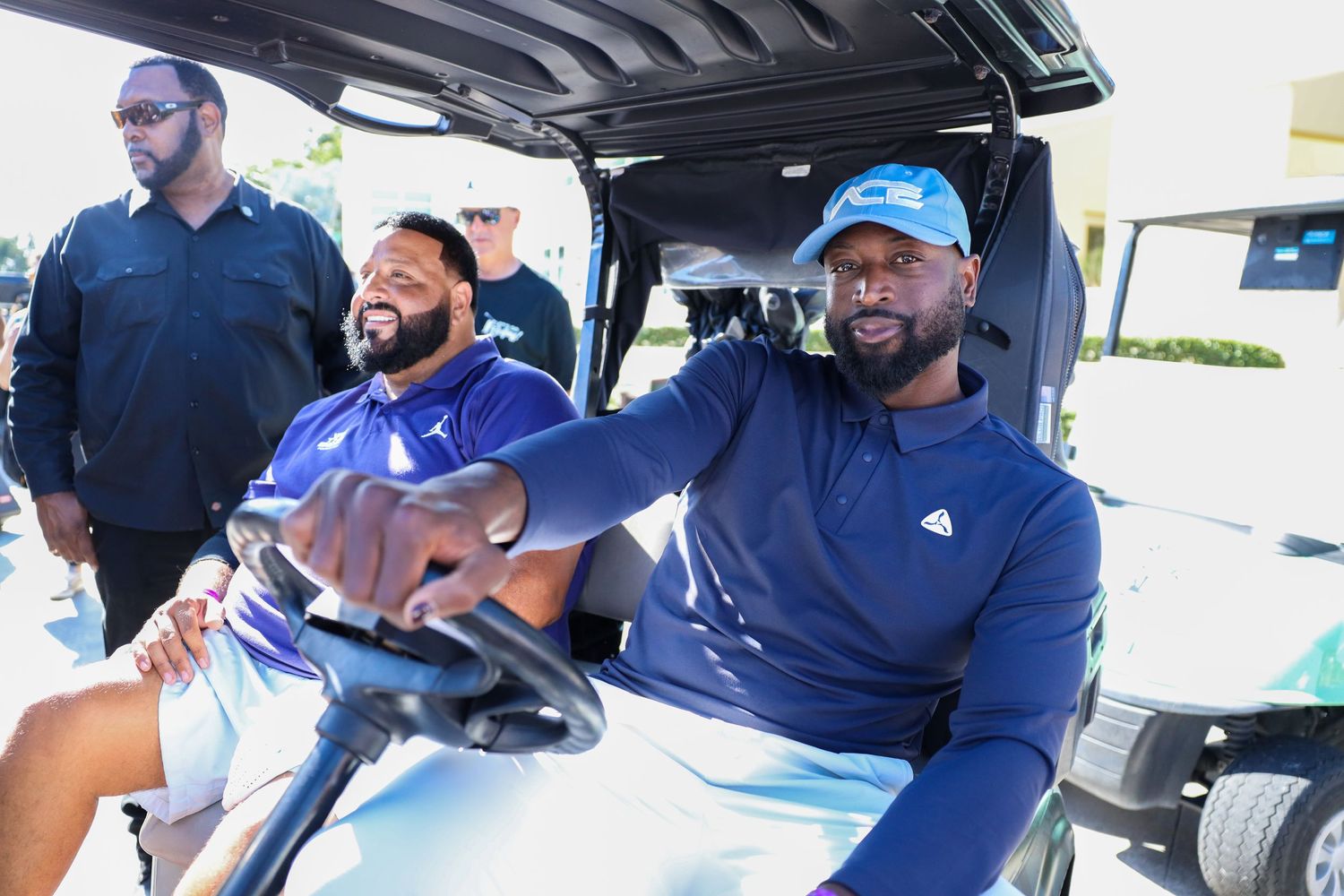 People seated in a golf cart wearing blue clothing and smiling during a sunny day outdoors.