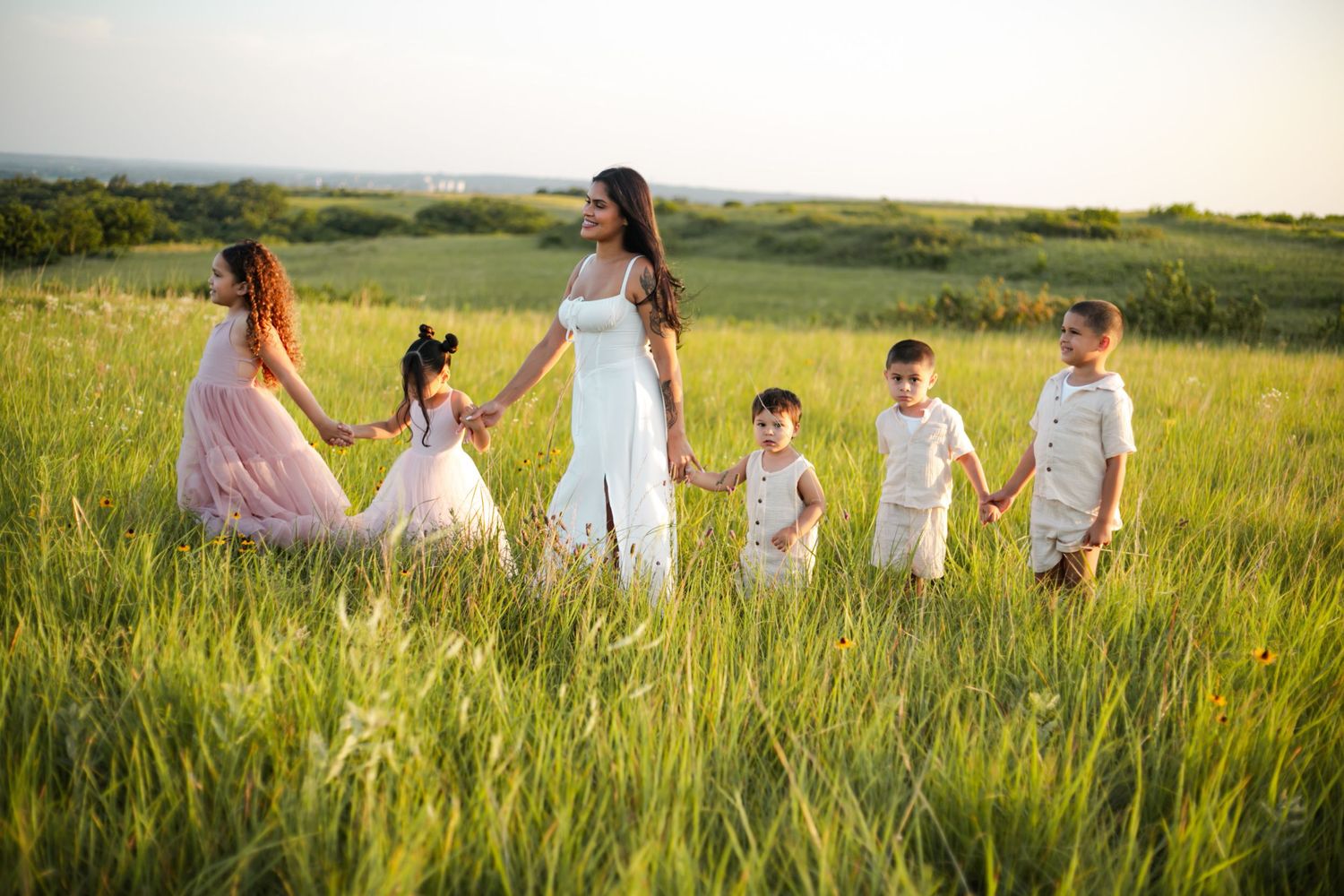 A group of people wearing flowy dresses and casual attire stand together outdoors during golden hour in a natural setting.