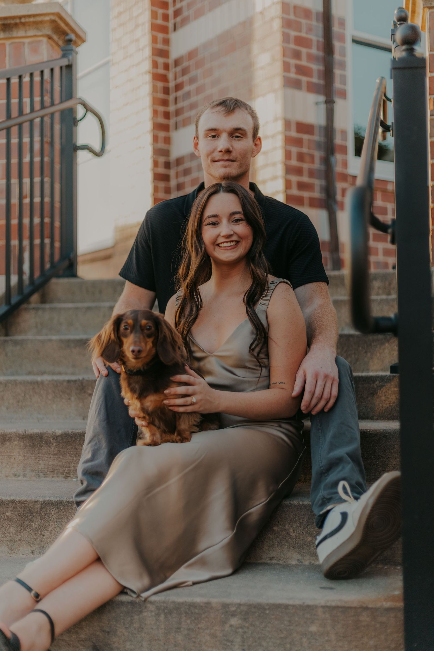 A couple sits with their dachshund puppy on brick steps during golden hour.