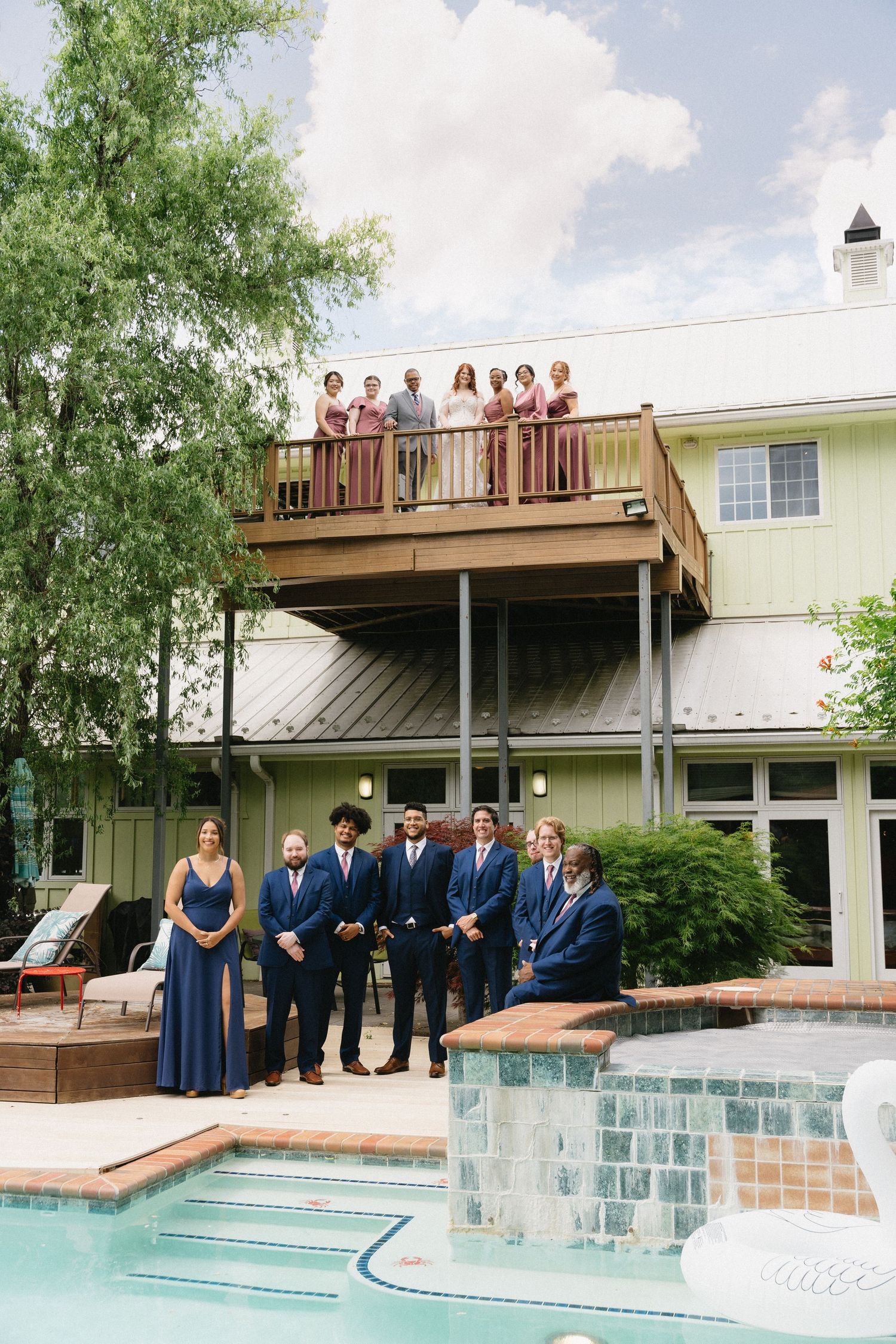 Wedding party poses by a pool with bridesmaids on balcony and groomsmen below.