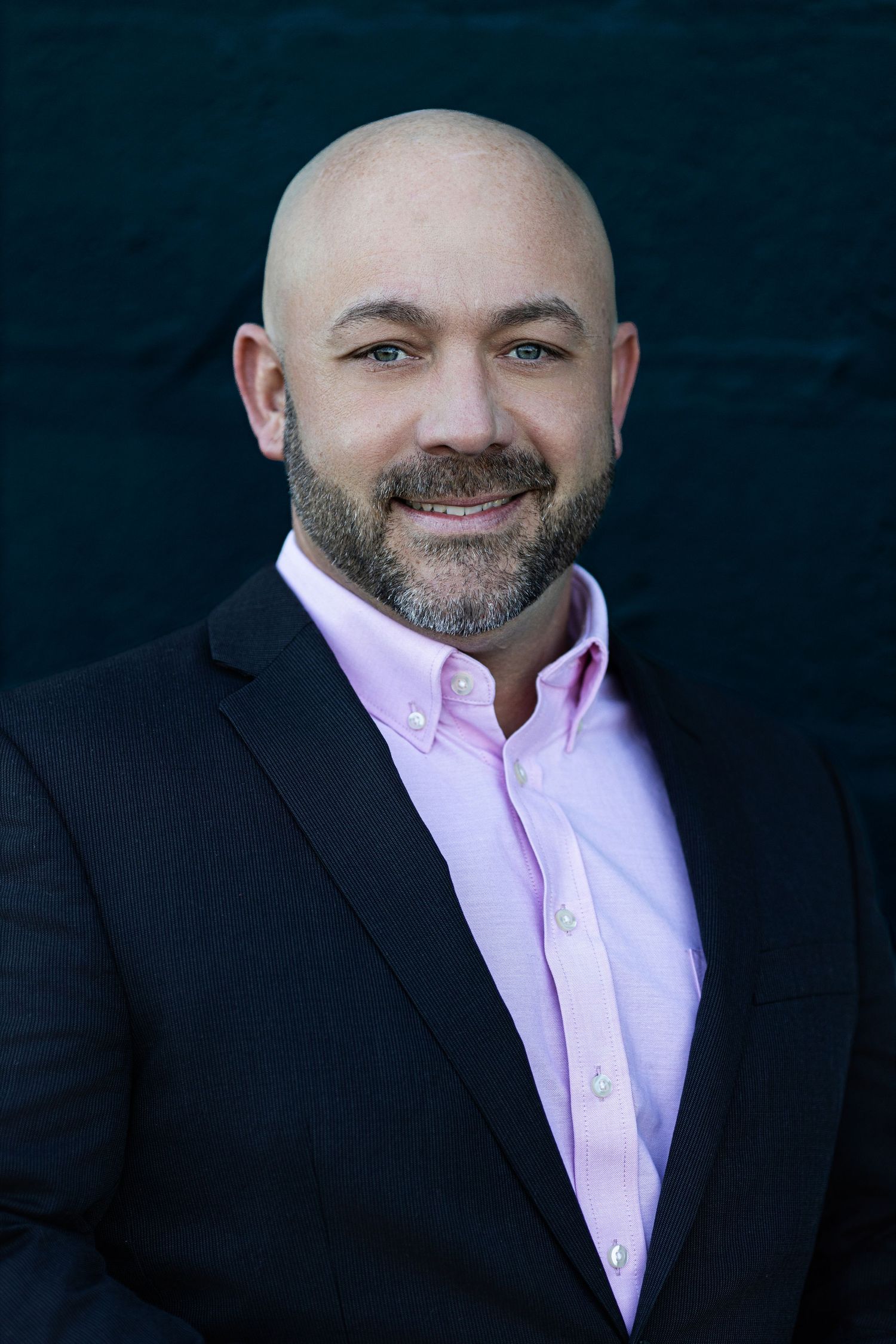 Professional business portrait of a person with a beard wearing a black suit jacket and pink button-down shirt against dark background.