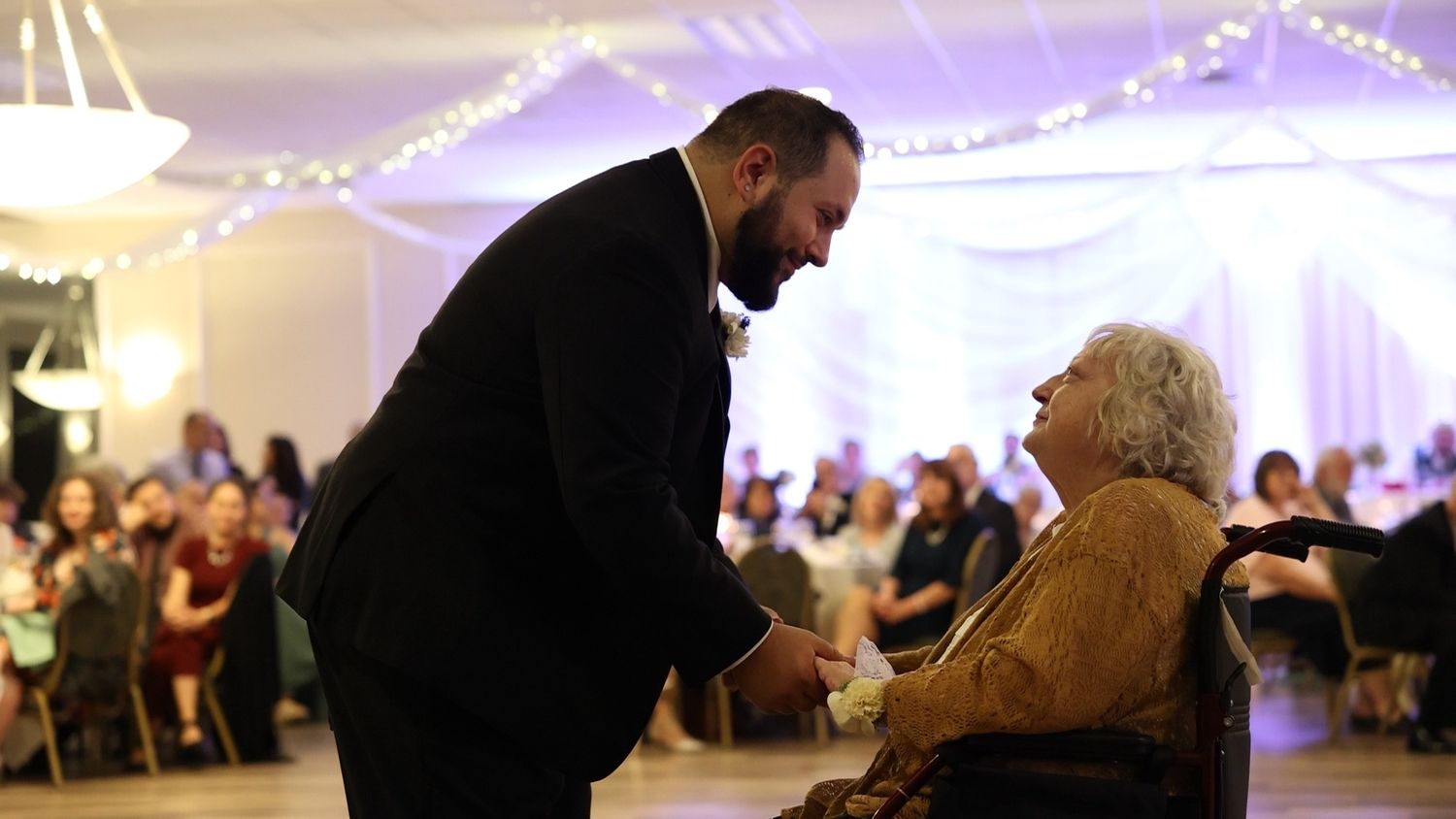 A person in dark suit bends down to greet an elderly individual in a wheelchair at an illuminated event venue.