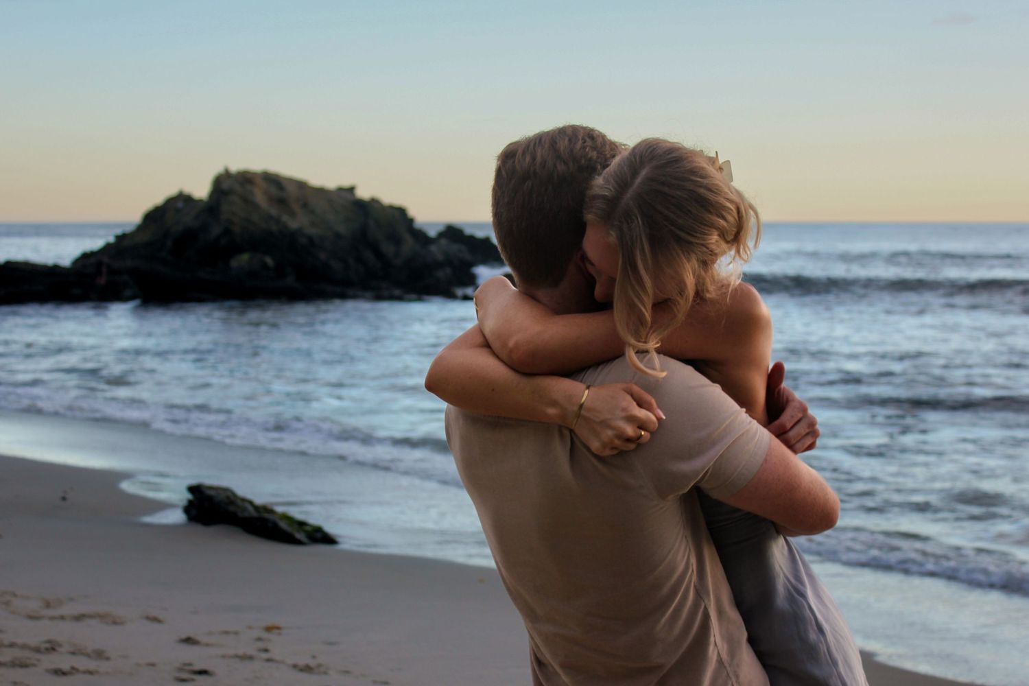A recently engaged couple hug on a beach with a rocky shoreline in a photography shoot. Waves crash upon the sand with a sunset background.
