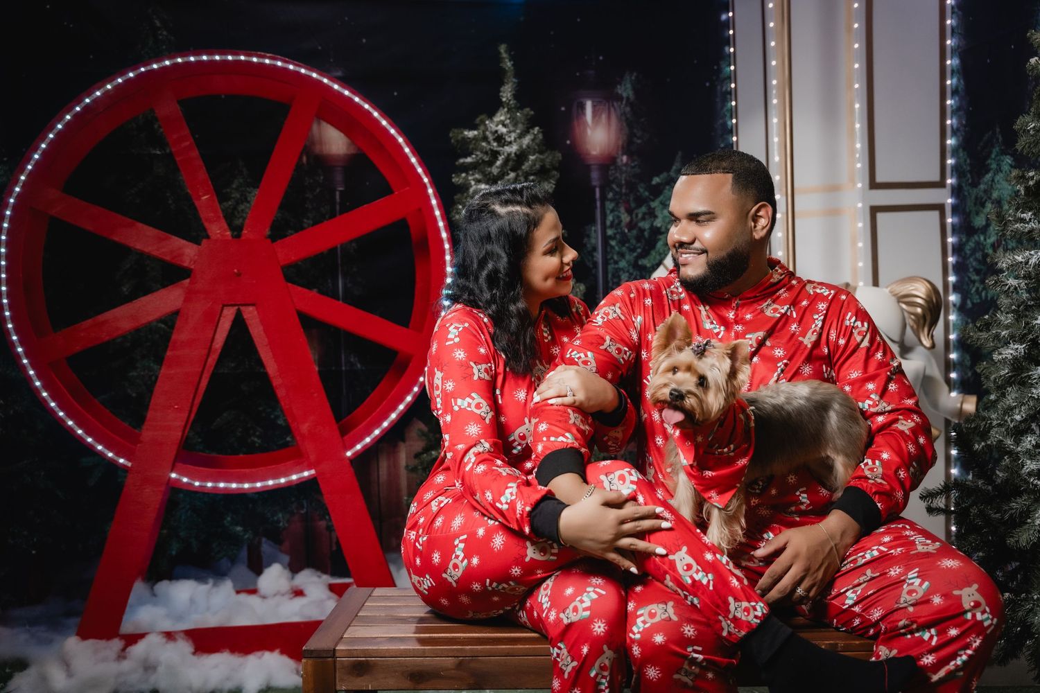 Couple in matching red Christmas pajamas pose with their dog by a decorative ferris wheel backdrop.