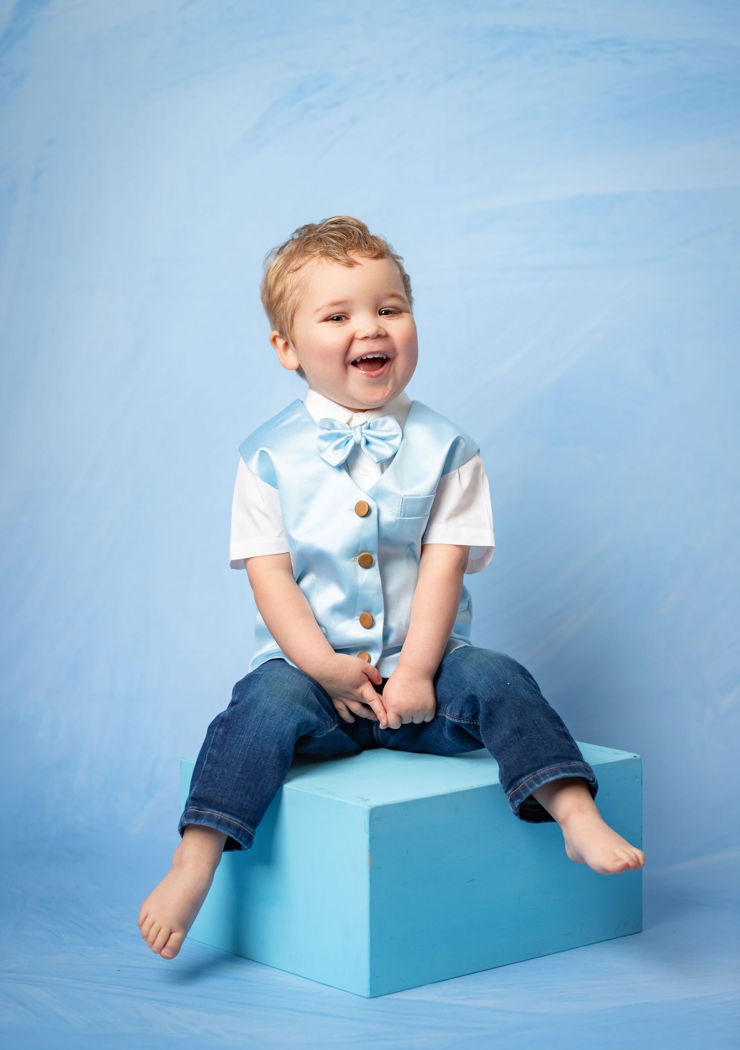 Portrait of smiling little one in light blue vest and jeans sitting on turquoise box against blue backdrop.