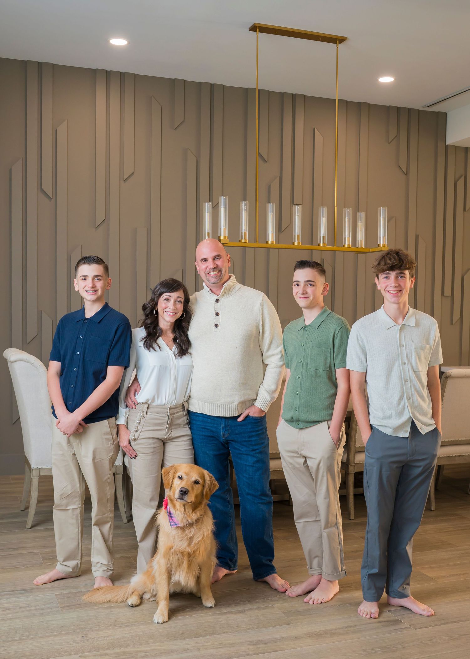 A casual indoor family portrait with a golden retriever dog in front of a modern accent wall and hanging light fixture.