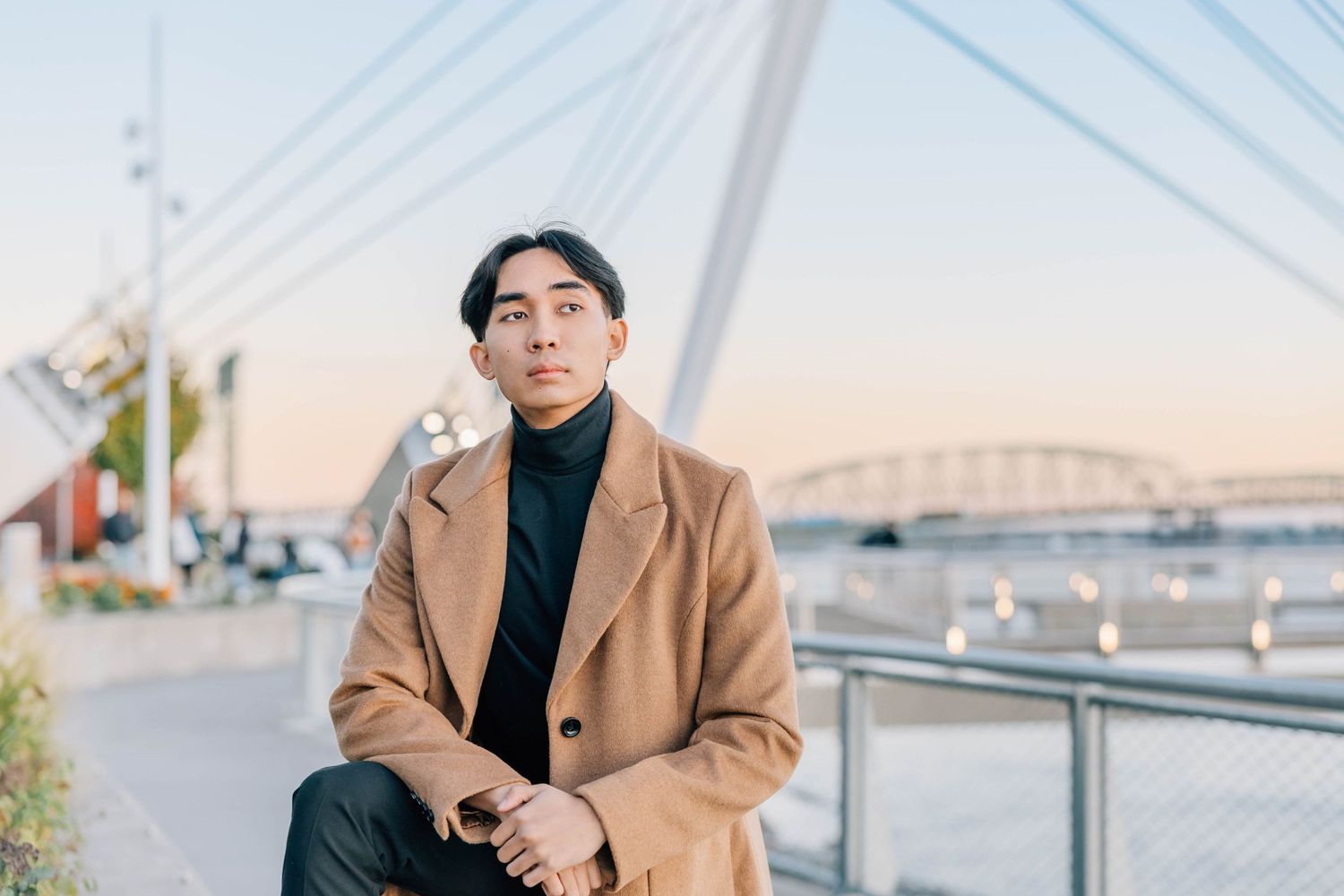 Young Asian man in camel coat sitting outdoors near waterfront bridge at golden hour sunset.