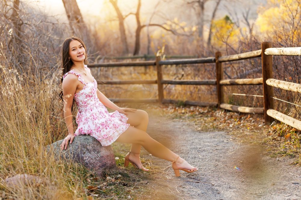 A person in a floral dress sits gracefully by a stream during golden hour in an autumn setting.