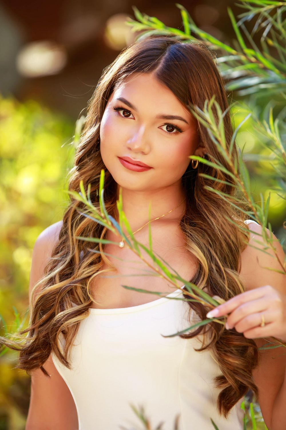 A portrait in nature featuring long wavy brown hair and a white top with green foliage in soft sunlit background.