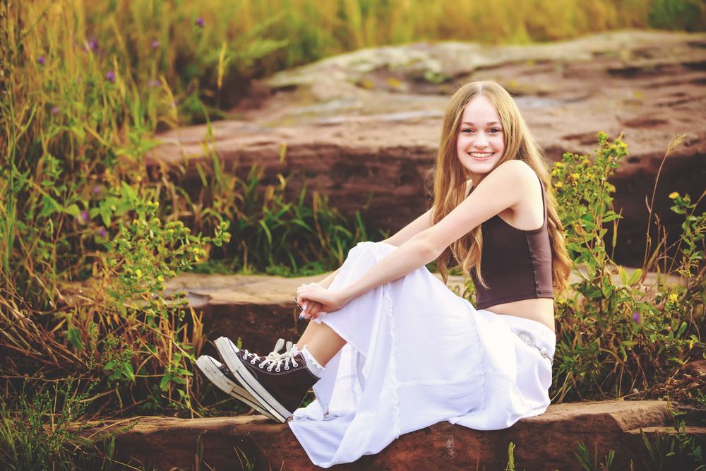 A person in a white dress and sneakers poses on stone steps surrounded by greenery at sunset.