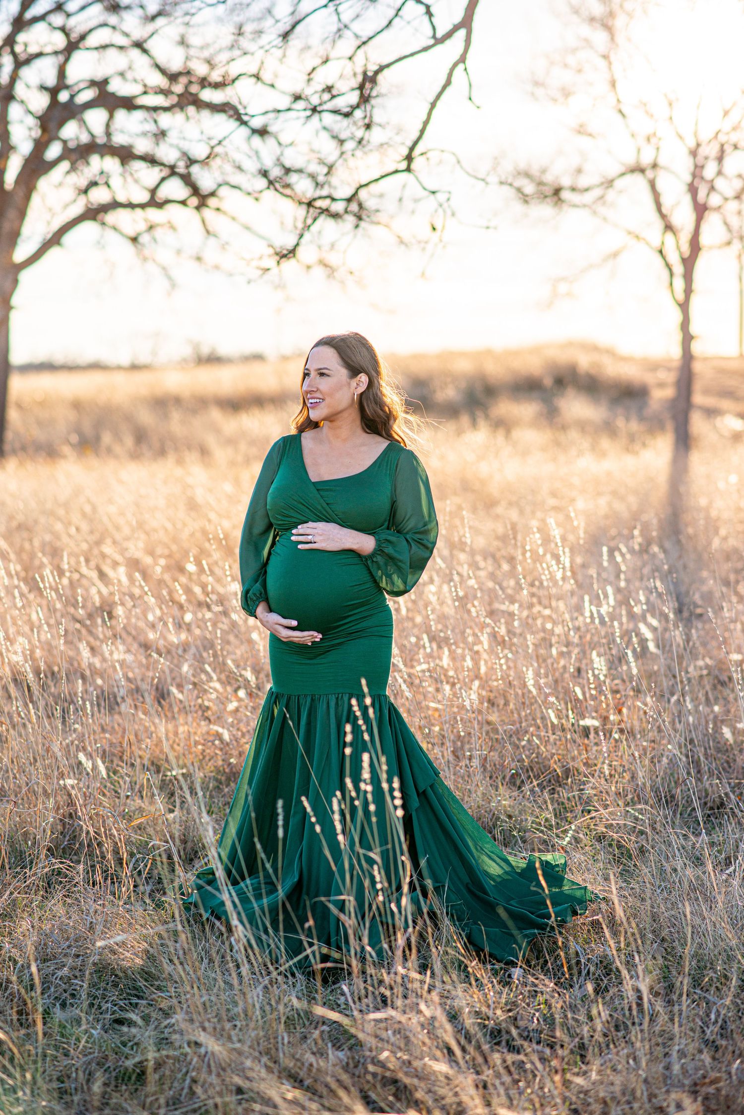 Expectant mother in flowing emerald green gown poses gracefully in golden field at sunset.