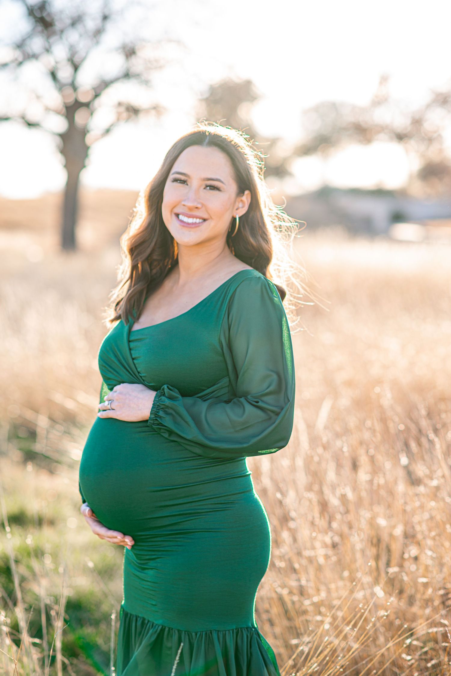 Glowing expectant mother in emerald dress posing in golden field at sunset.