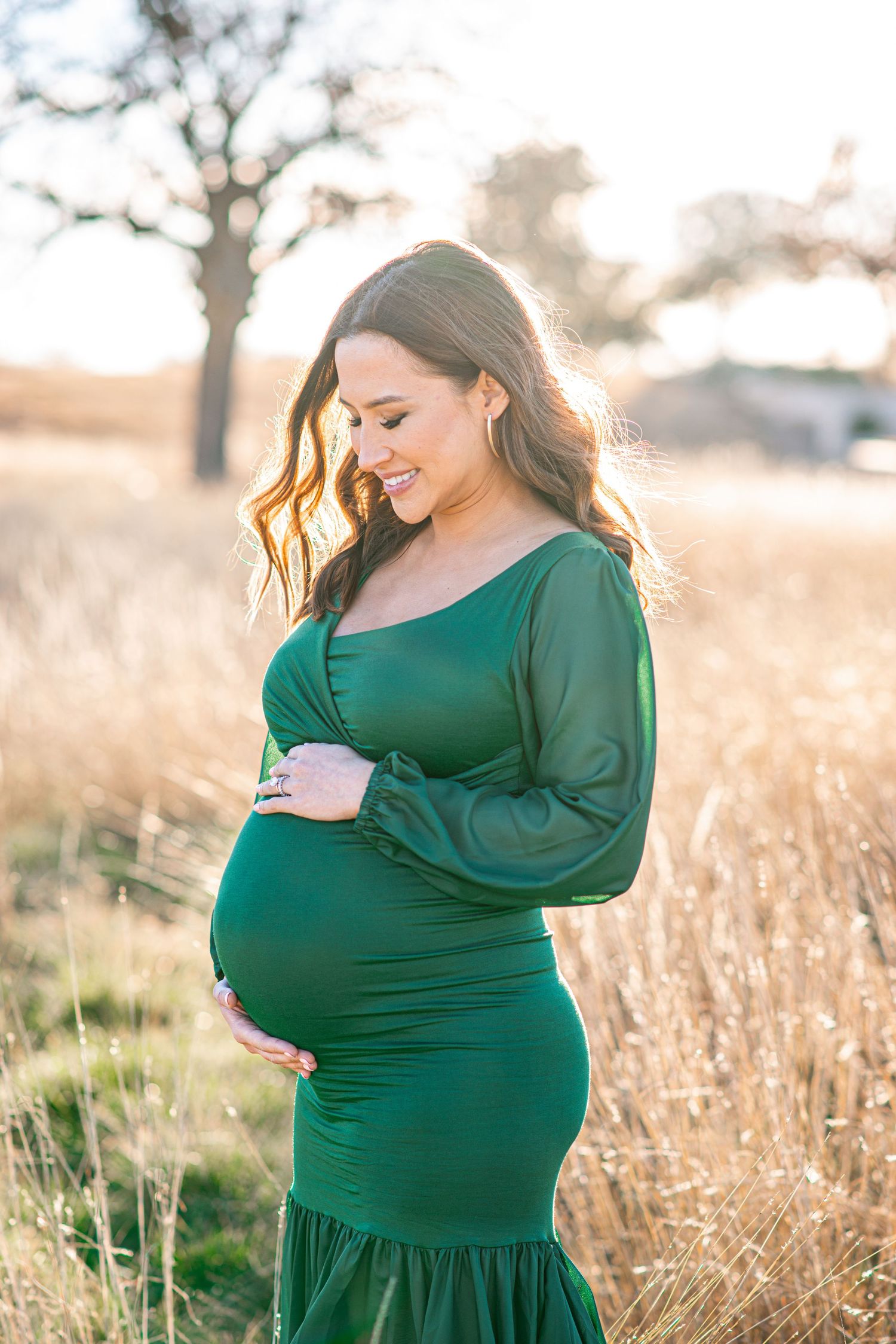 A maternity photo of someone in an elegant emerald green dress standing in a golden field at sunset.