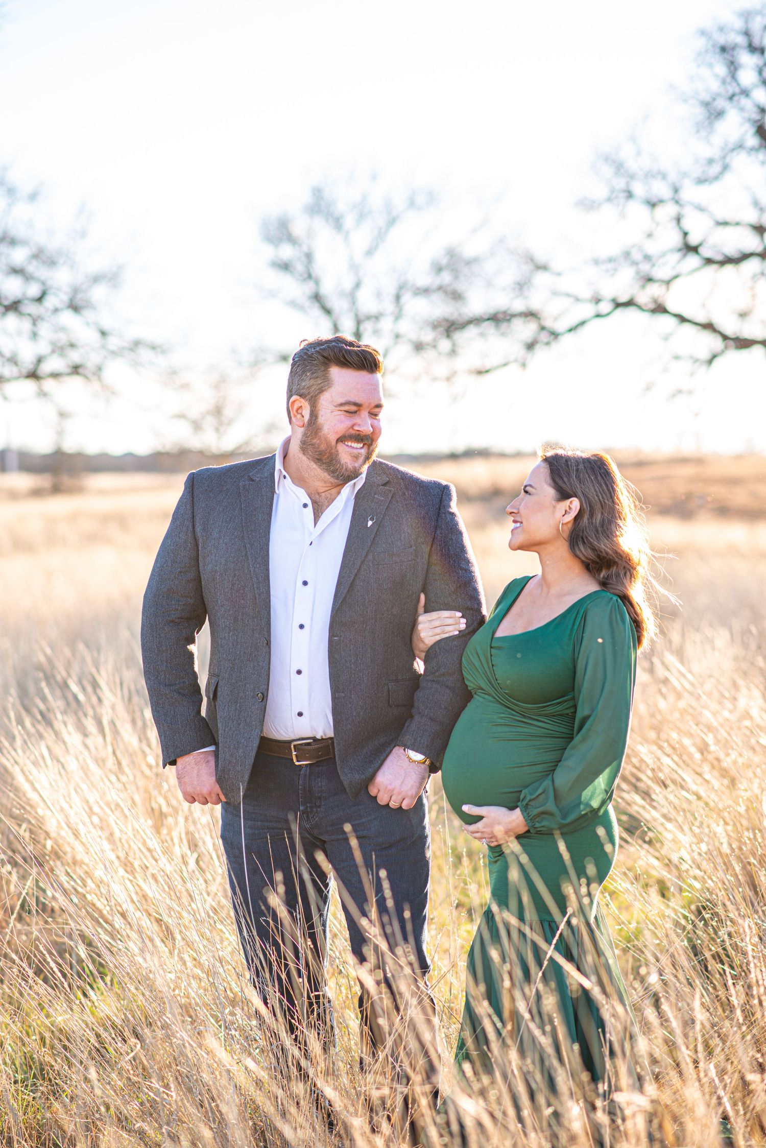 Couple in formal attire share intimate moment during maternity photoshoot in golden field.