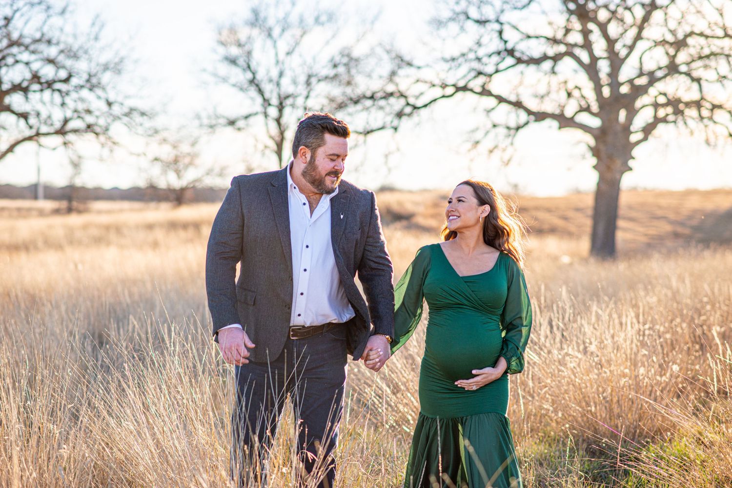 Joyful couple walking through golden field during sunset maternity session.