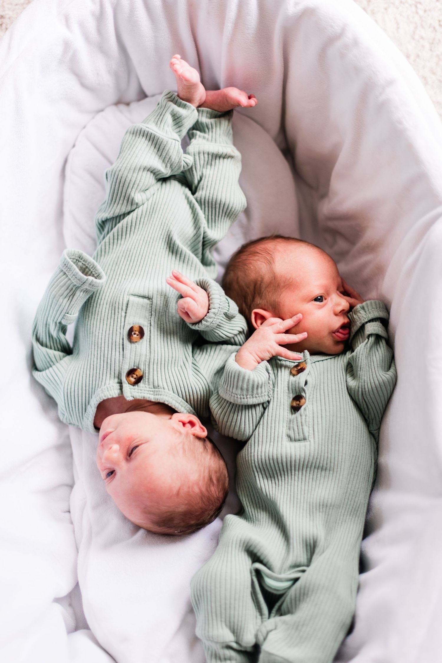 Two infants in matching sage green button-up onesies lying in white bassinet.