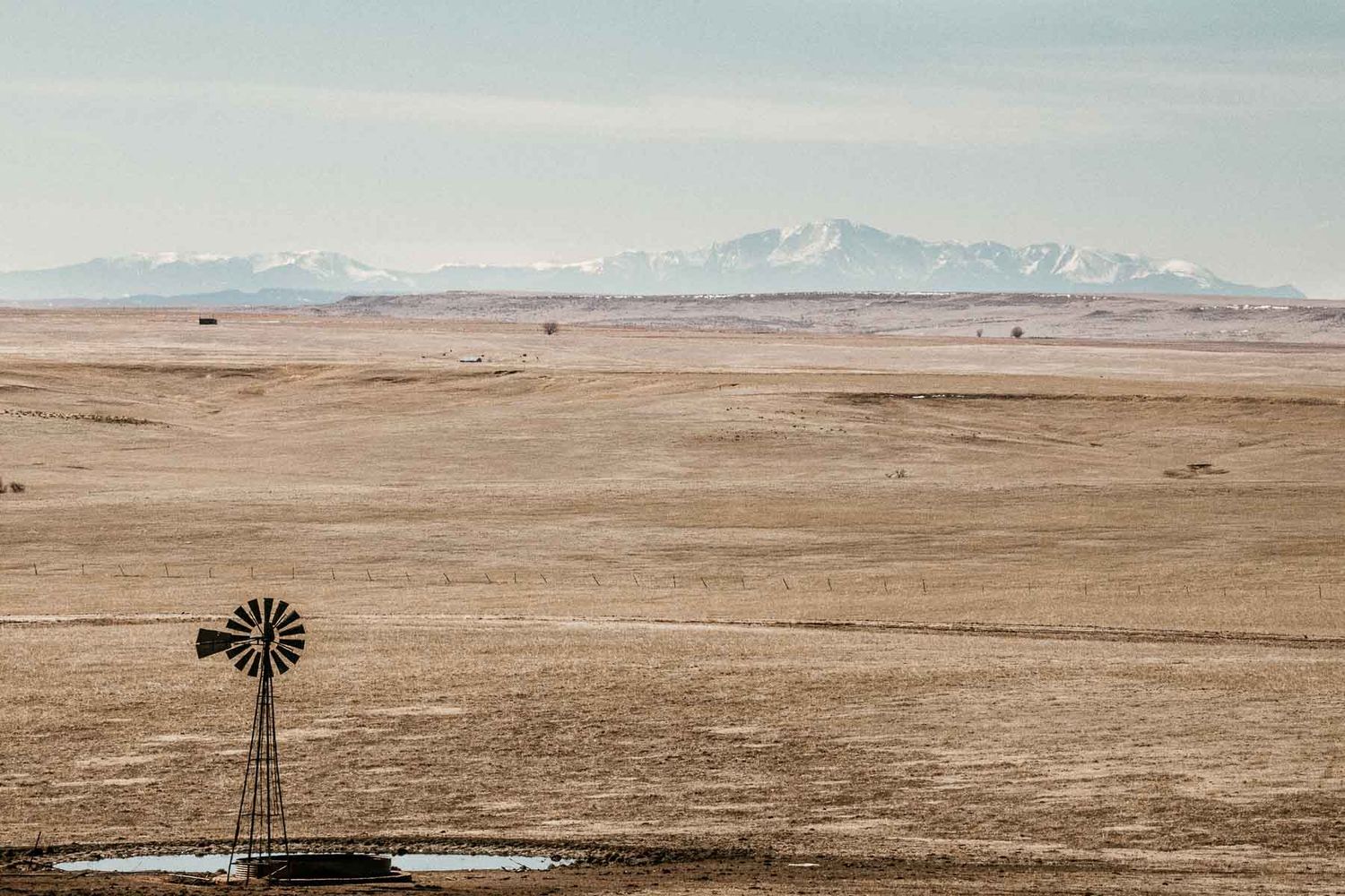 A lone weather vane on the Colorado plains at the foot of the Rocky Mountains, with the mountain peaks visible in the distance.