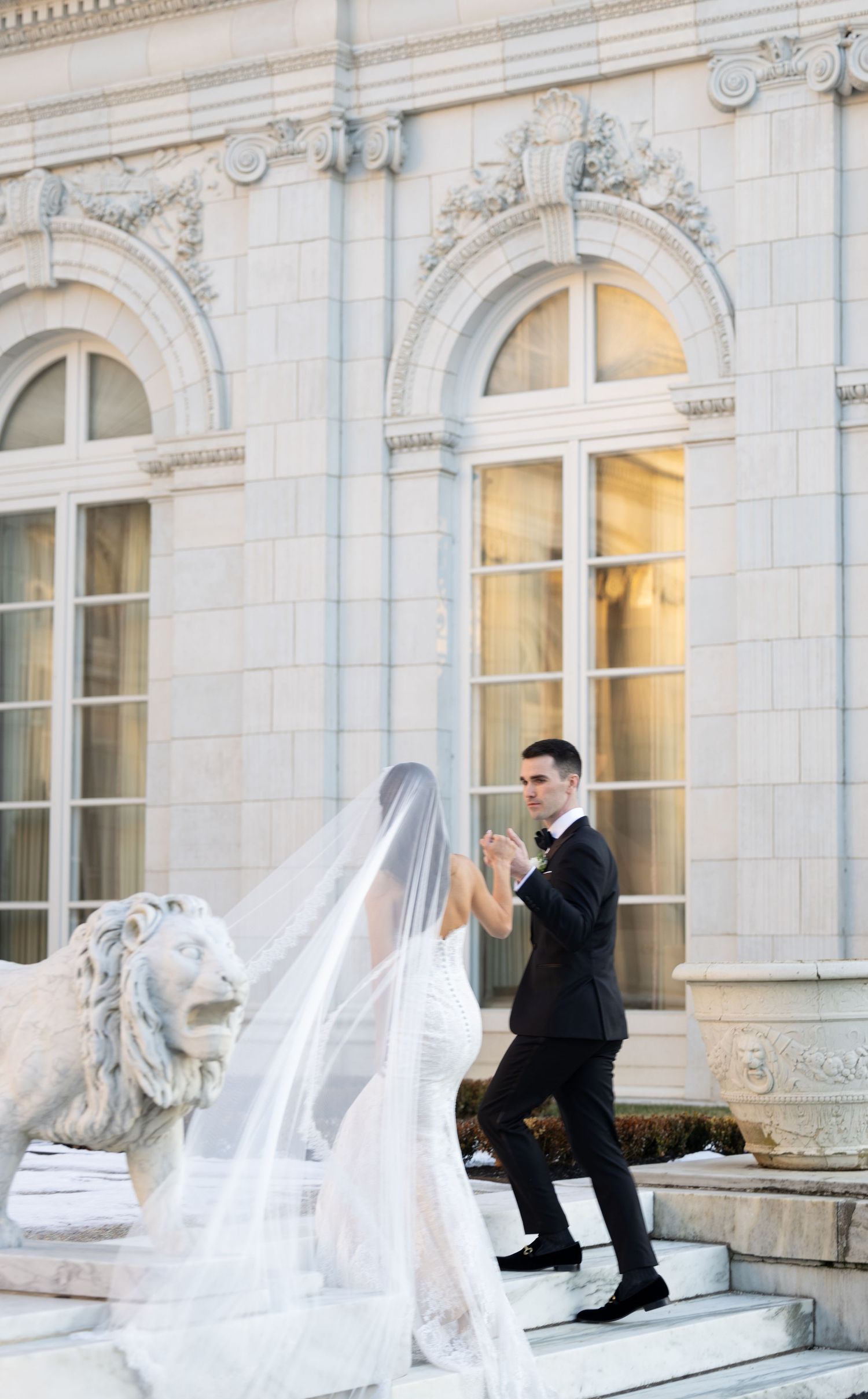 A wedding couple poses on grand marble steps of an elegant historic building with ornate architectural details and lion statue.