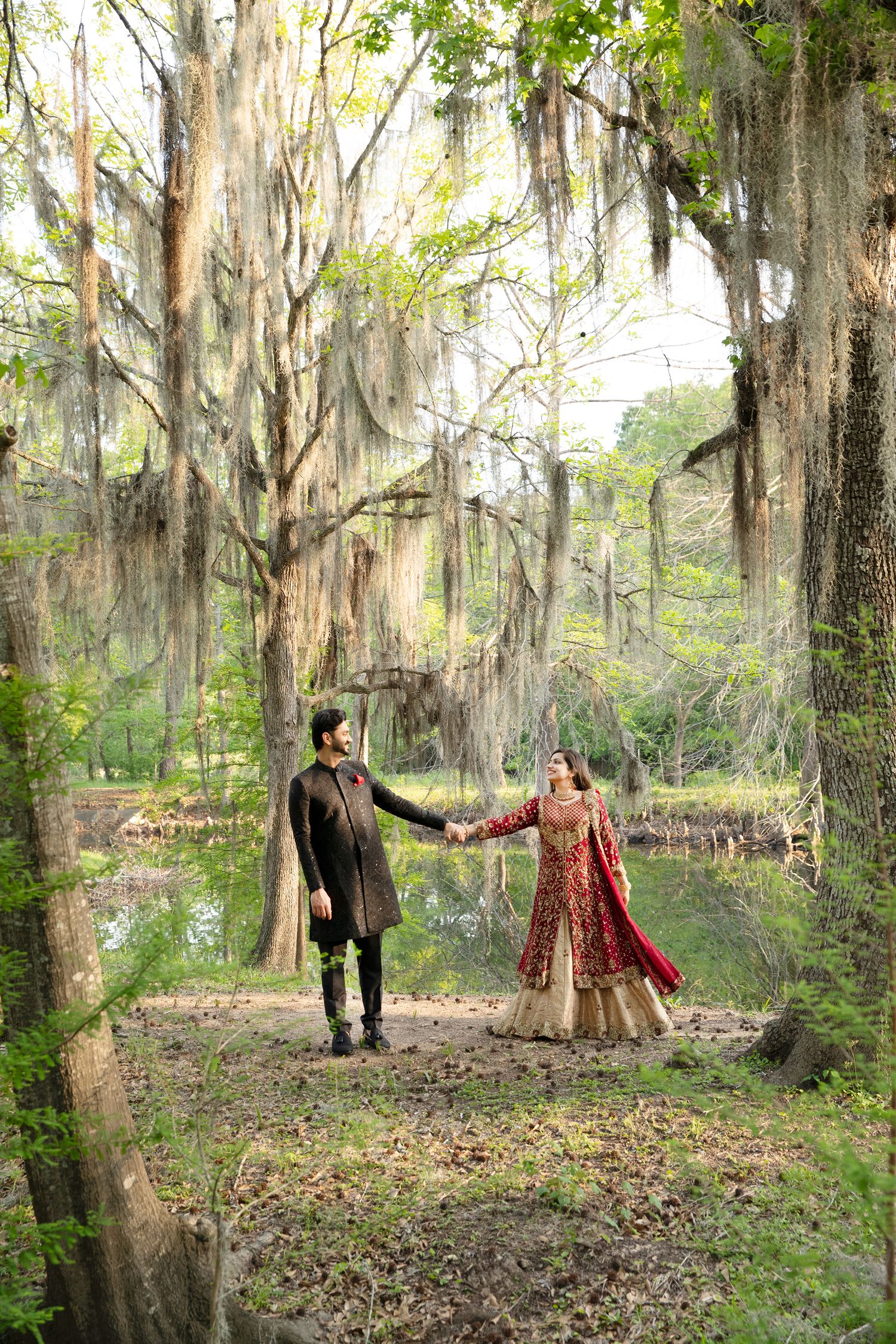A couple in traditional South Asian attire hold hands in a dreamy Spanish moss-covered forest setting during a photoshoot.