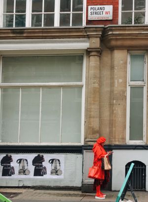 A person in a bright red coat stands on Poland Street in London against a building with poster-covered windows.