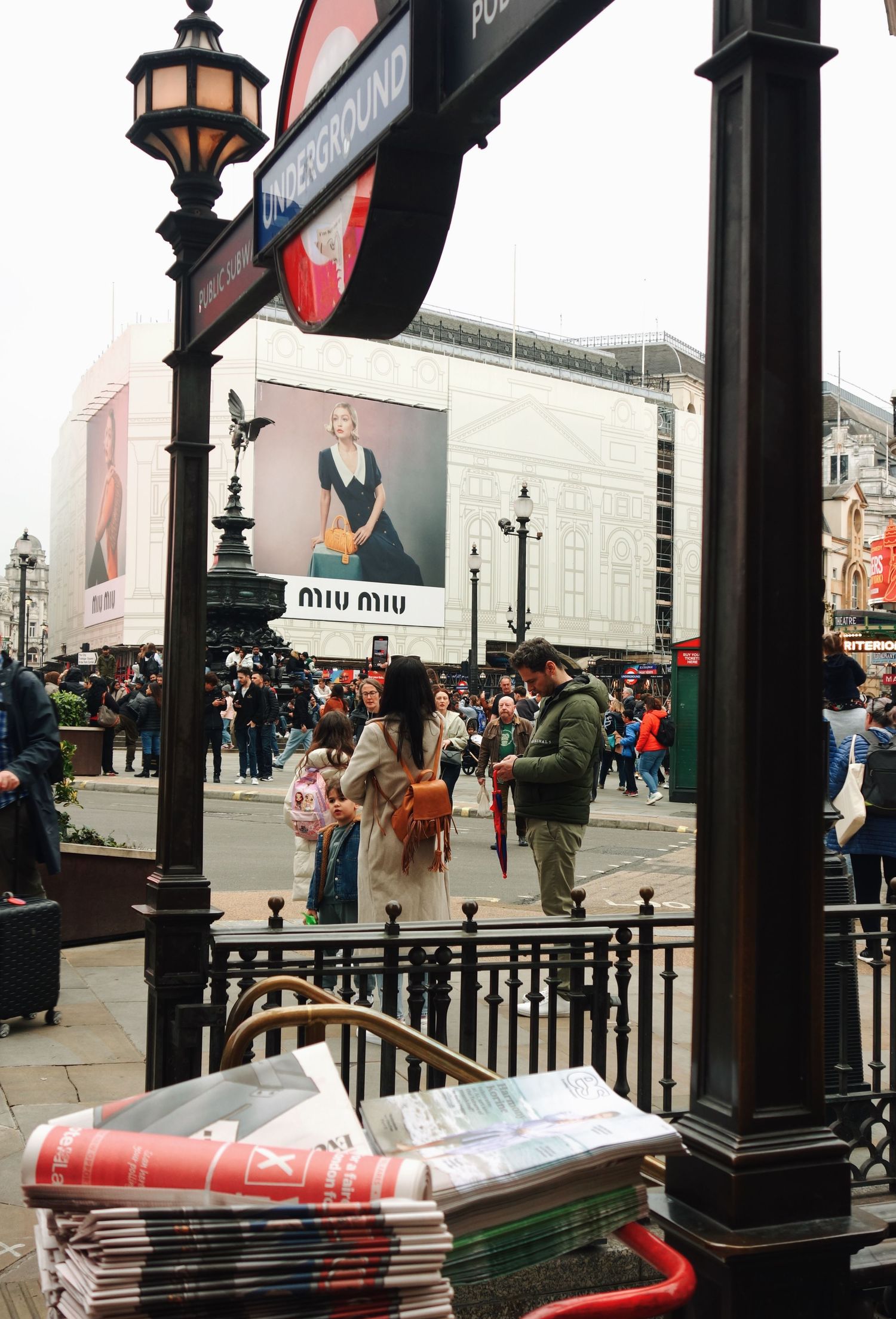 Busy street scene outside Piccadilly Circus underground station in London with Miu Miu advertisement billboard and ornate lamppost.