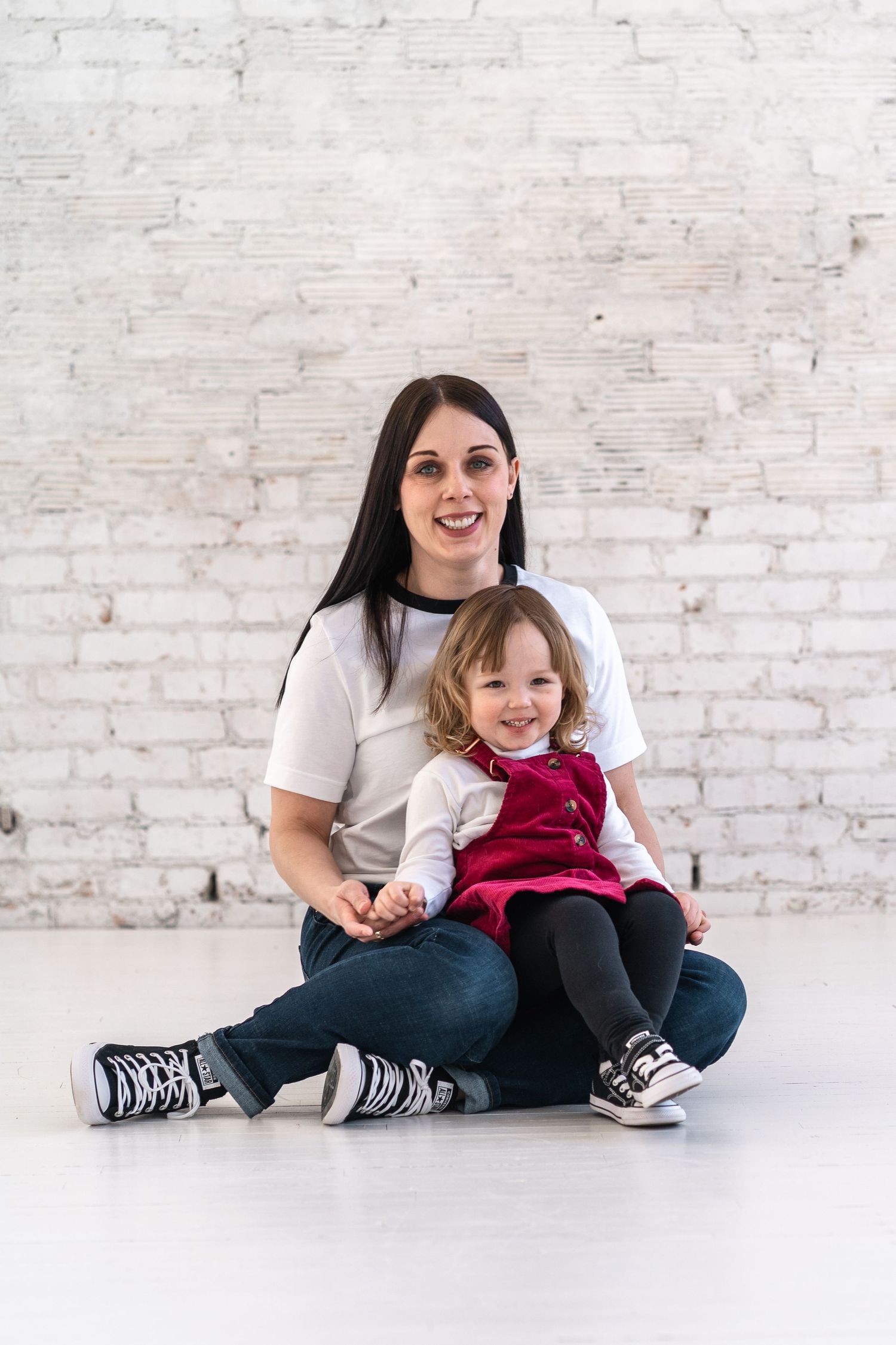 Two people sitting together on floor against white brick wall wearing casual outfits.
