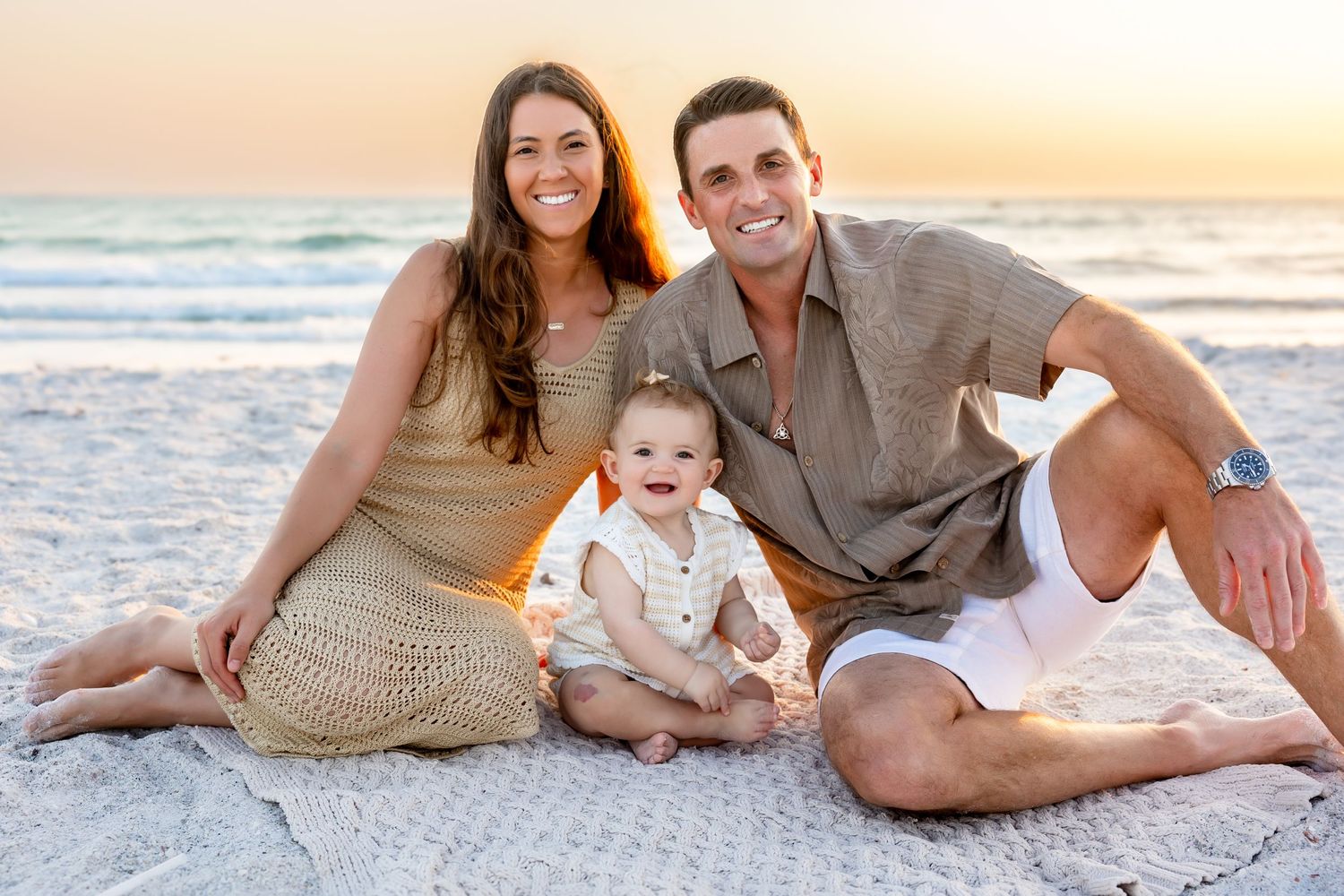 Happy family with parents and baby sitting together on sandy beach at sunset.